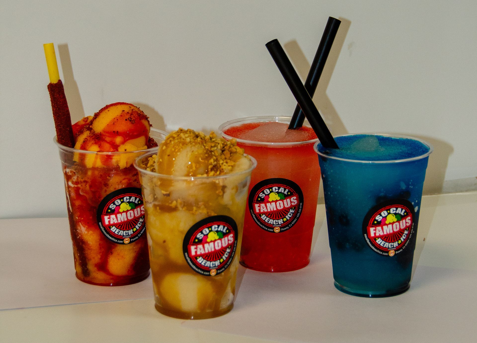 Four plastic cups containing colorful, slushy-style Mexican snacks and drinks, each branded with a circular logo.
