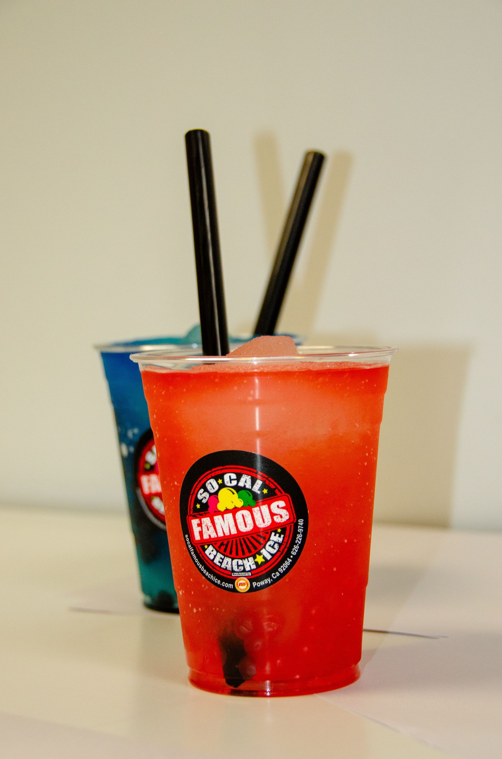 Two plastic cups filled with brightly colored red and blue frozen drinks, each with a black straw, against a plain wall.