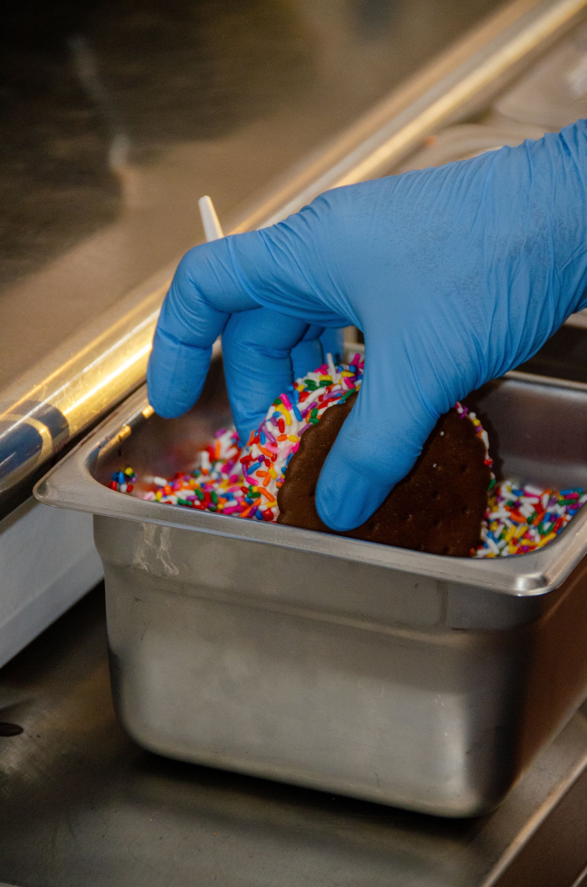 A gloved hand dips a chocolate cookie into a metal tray filled with colorful rainbow sprinkles.