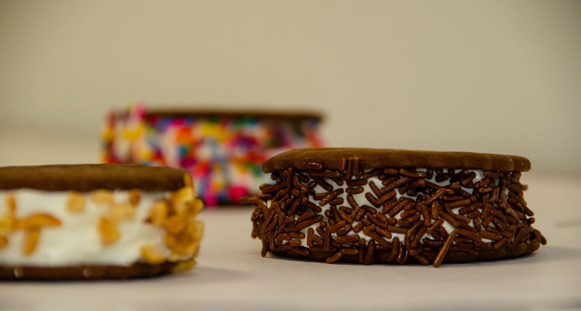 Three ice cream sandwiches topped with different sprinkles sit on a white surface.