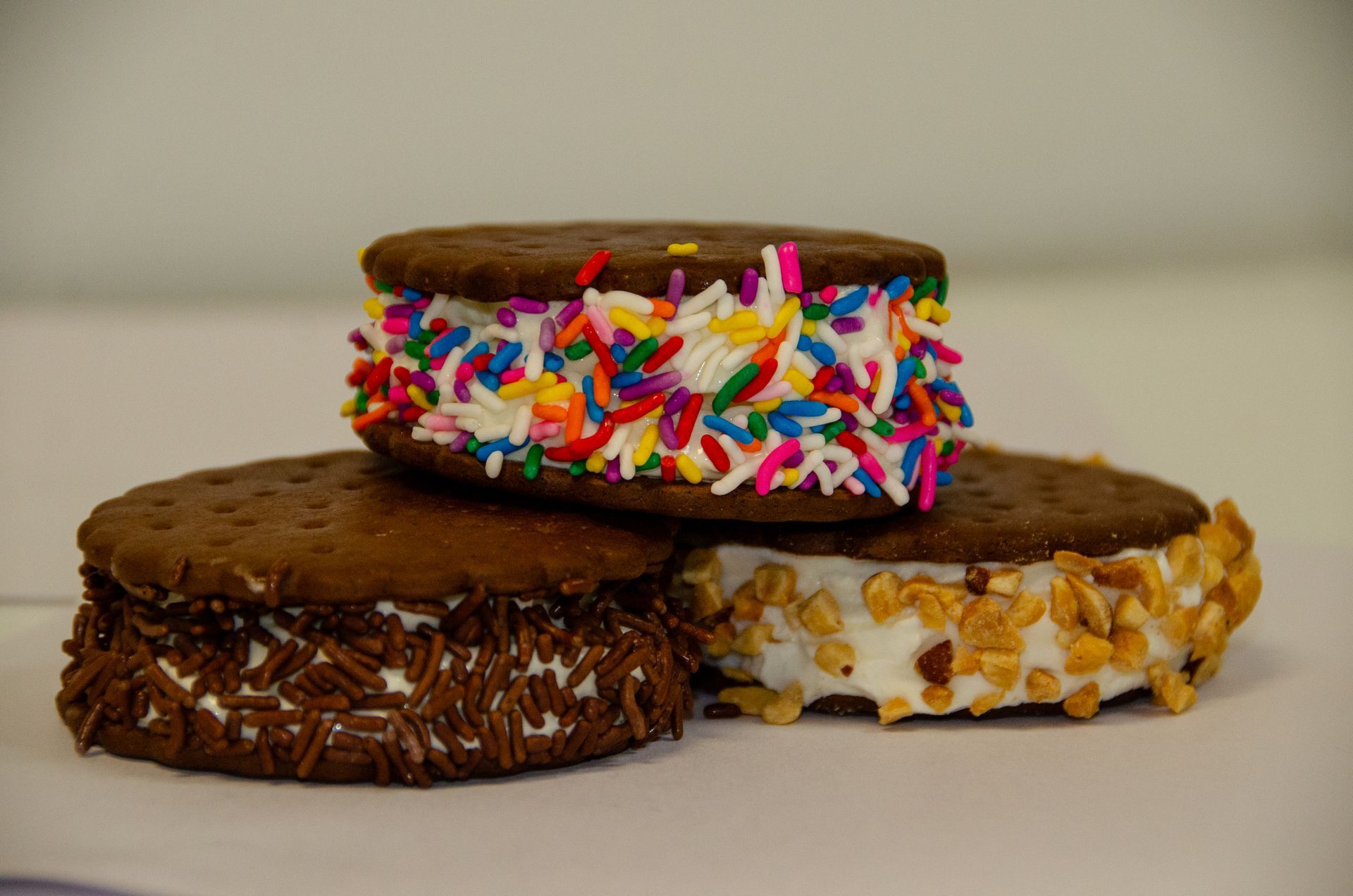 Three ice cream sandwiches topped with rainbow sprinkles, chocolate sprinkles, and chopped nuts against a white background.