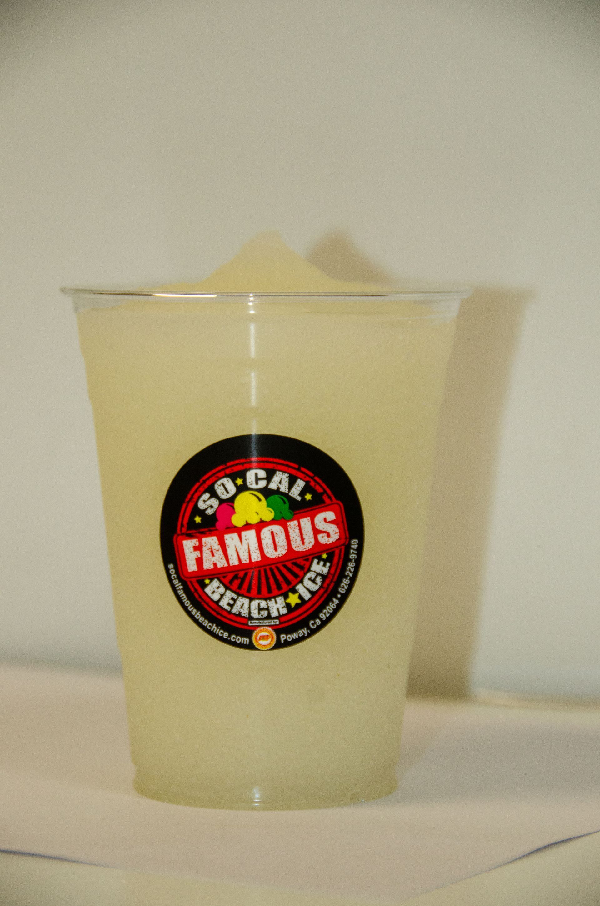 A clear plastic cup of pale yellow shaved ice with a So-Cal Famous Beach Ice logo sticker, centered against a light wall.