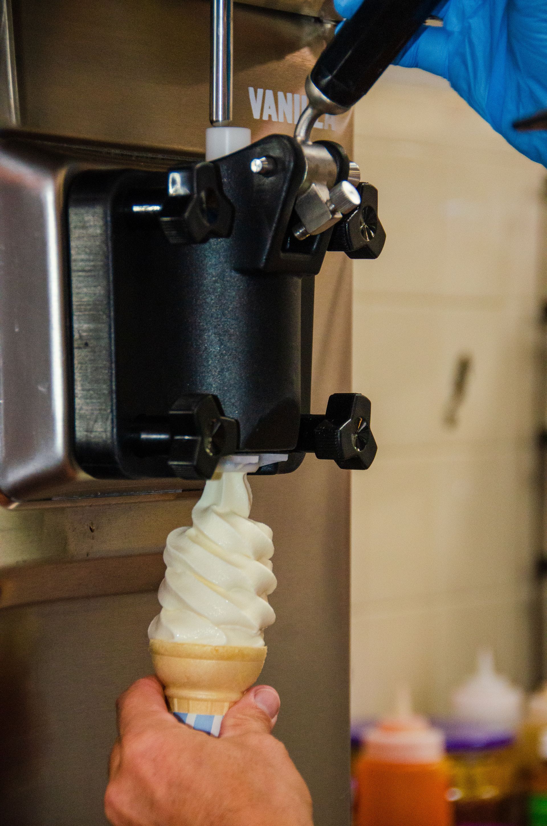 A hand in a blue glove operates a machine dispensing a vanilla soft-serve ice cream cone.