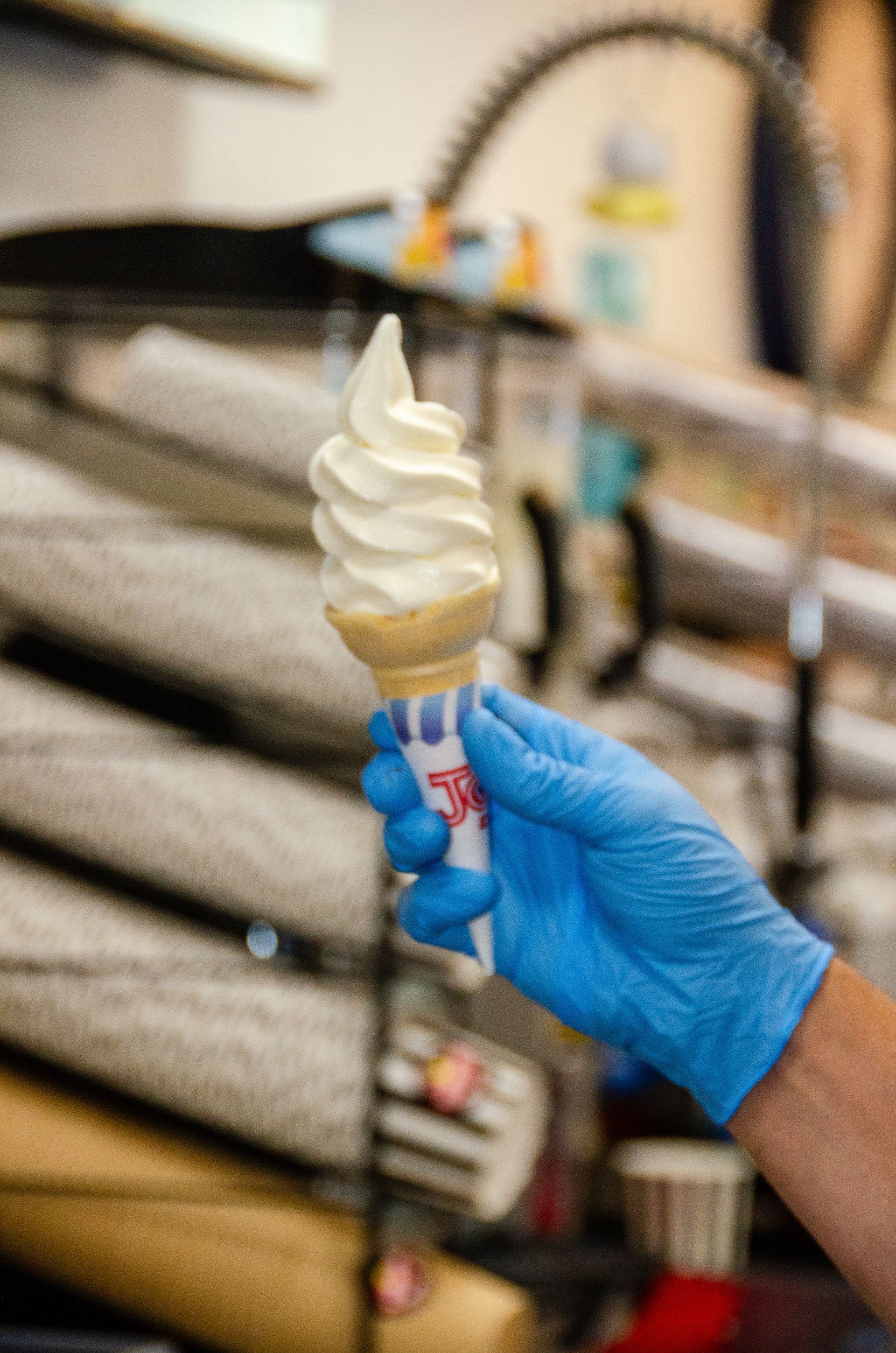 A hand wearing a blue latex glove holds a vanilla soft-serve ice cream cone in a commercial kitchen setting.