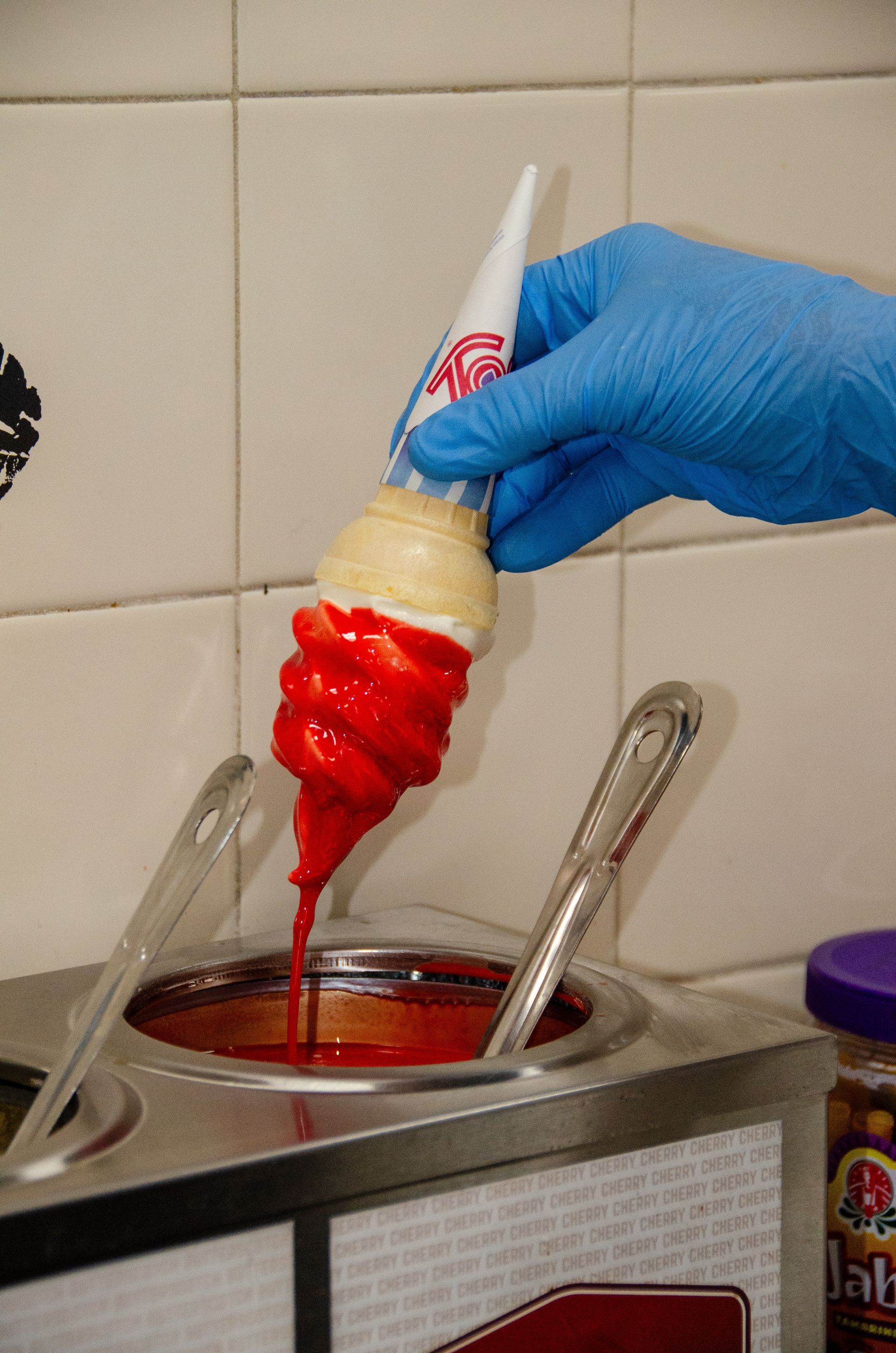 A gloved hand dips a soft-serve ice cream cone into a container of bright red liquid coating.