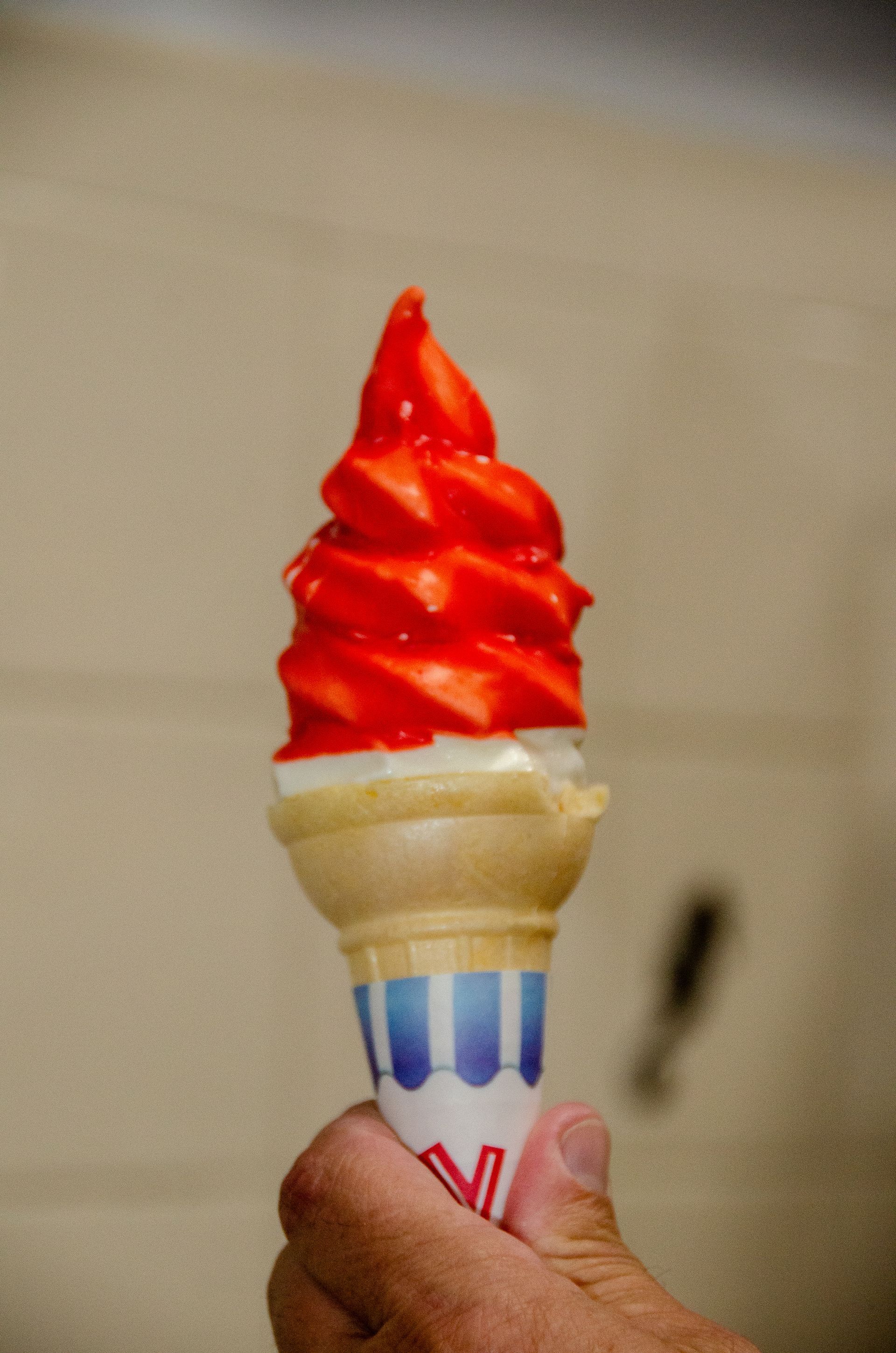 A hand holds a soft-serve ice cream cone topped with a bright red, spiraled coating against a plain wall.