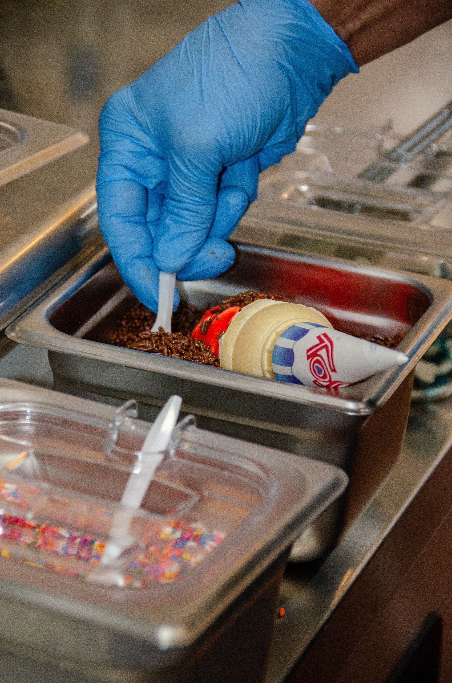 A gloved hand dips an ice cream cone into a container of sprinkles in a food prep area.