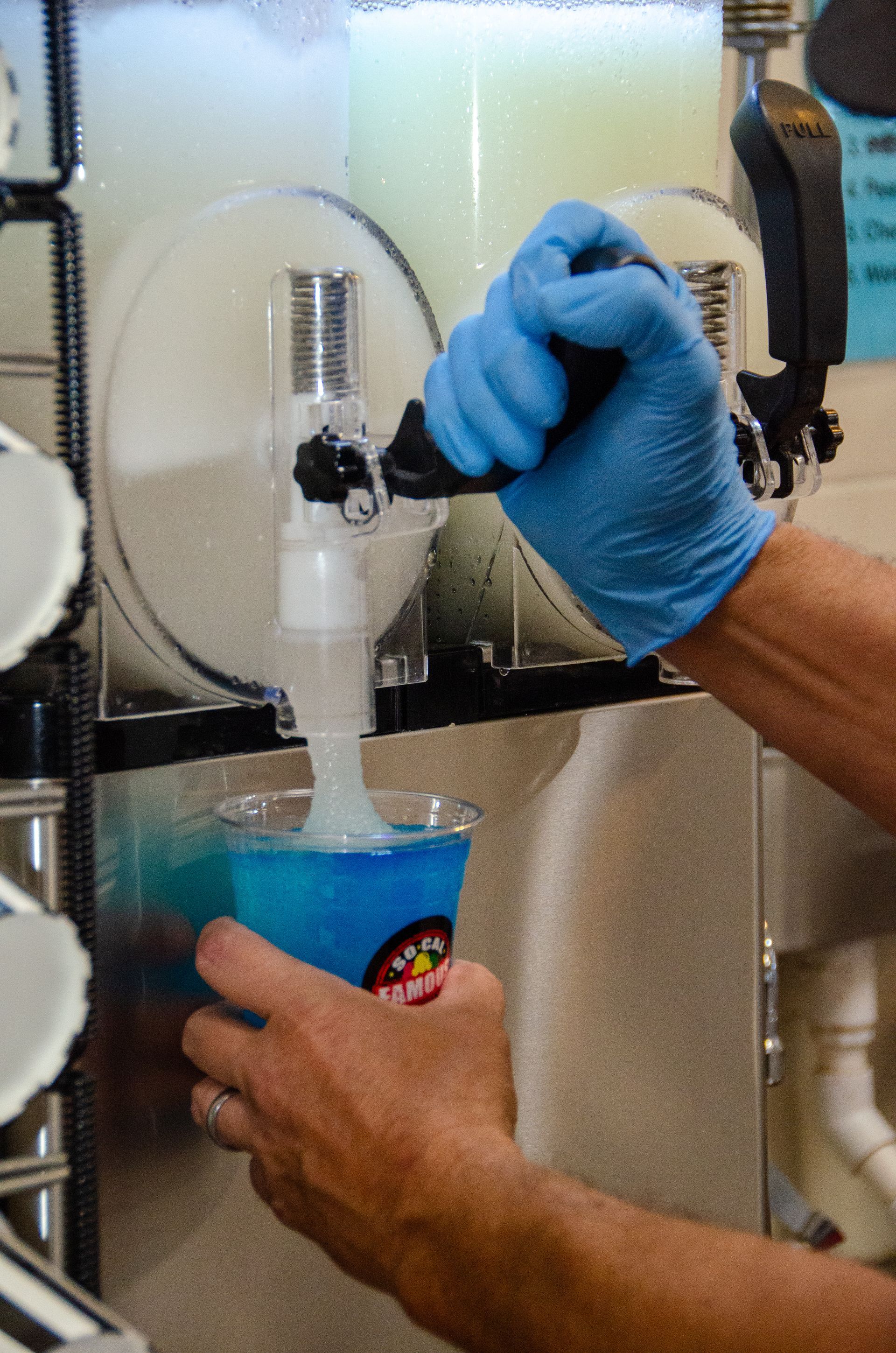 A gloved hand pulls a lever on a slushie machine, dispensing a bright blue frozen drink into a clear plastic cup.