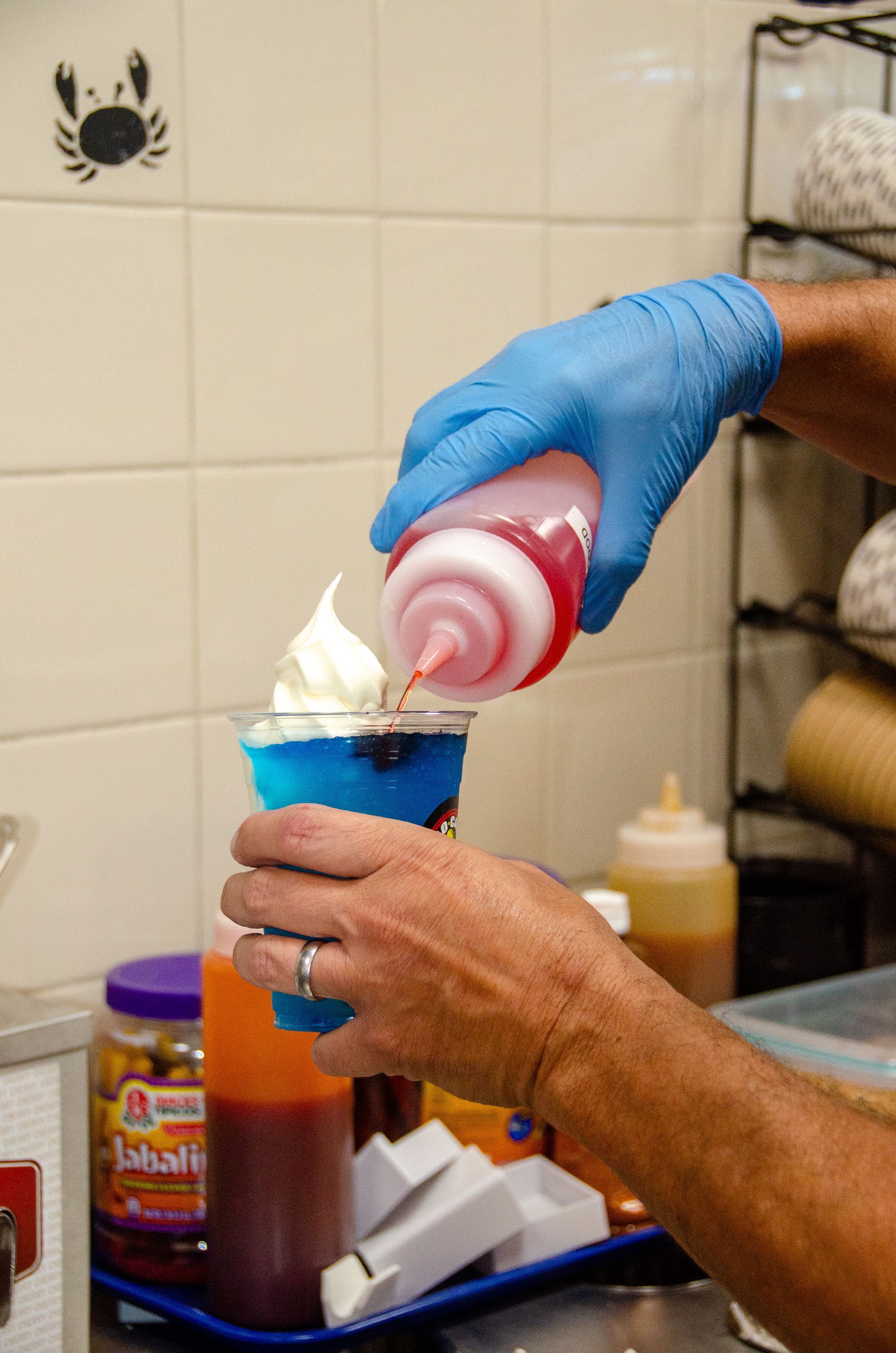 A gloved hand squeezes a red syrup bottle over a blue slushy topped with white soft-serve in a restaurant kitchen.
