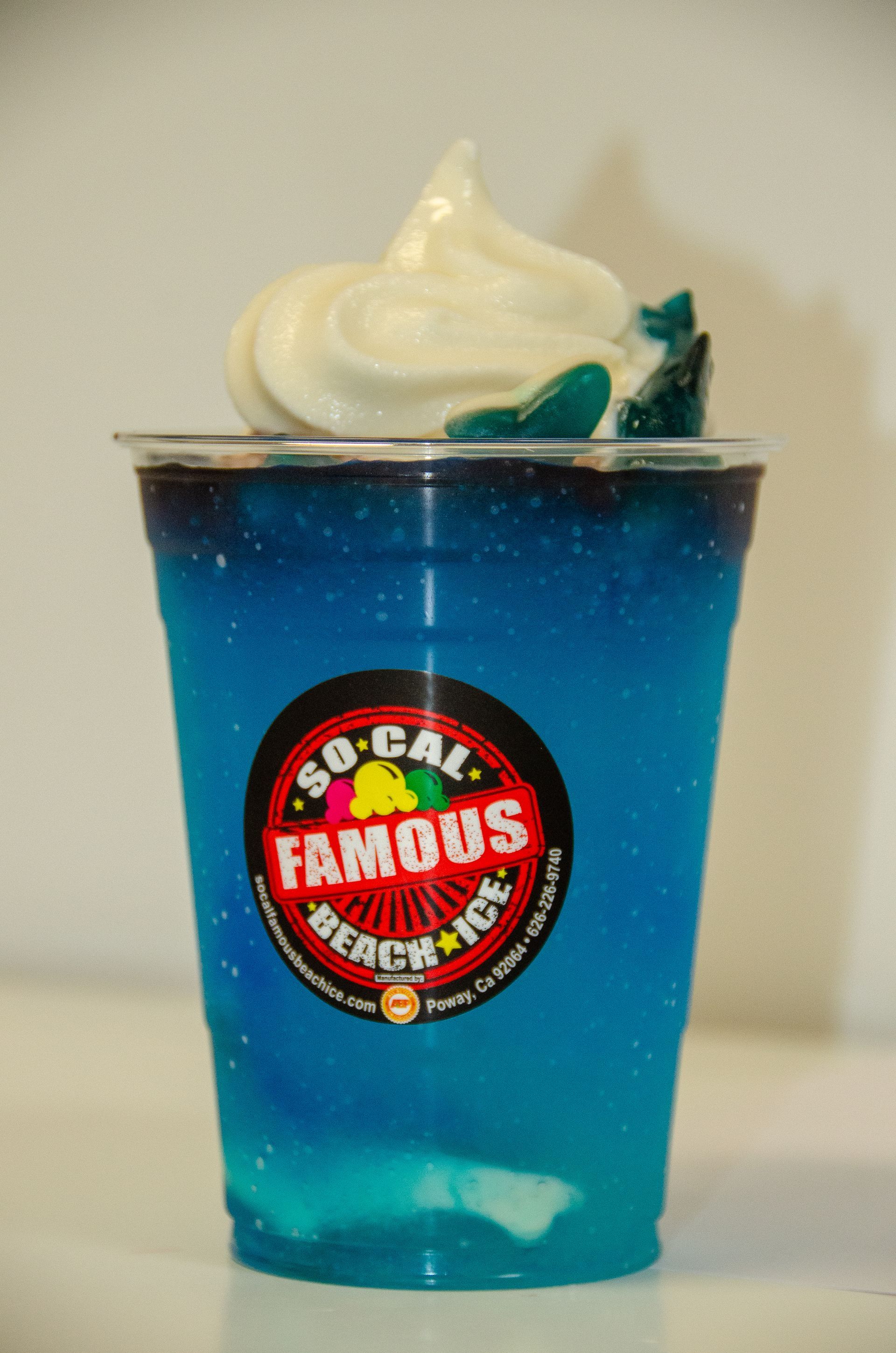 A plastic cup of bright blue shaved ice topped with white soft-serve and a gummy shark, featuring a So-Cal Beach Ice logo.