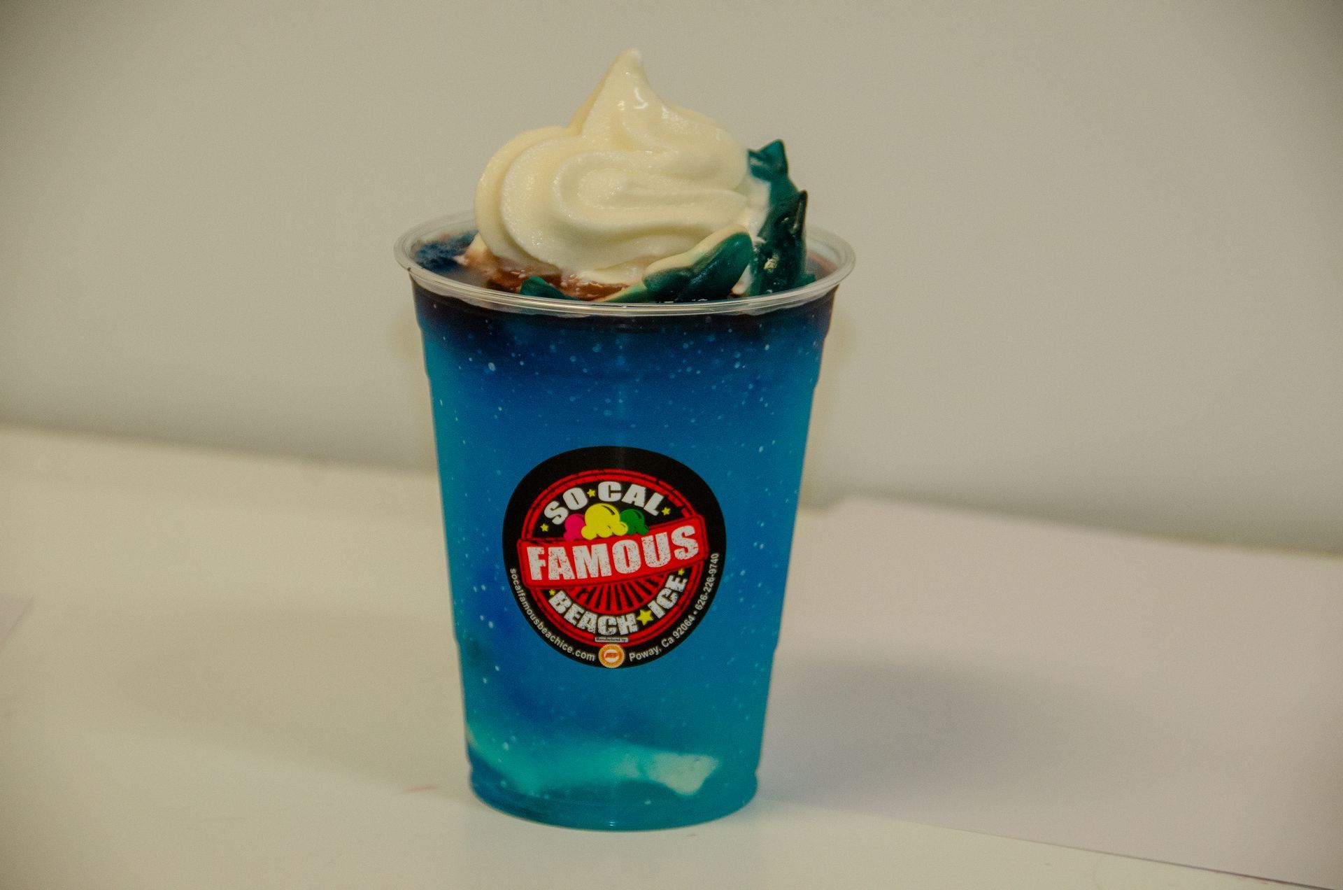 A clear plastic cup of bright blue slushy topped with a swirl of white soft-serve ice cream, featuring a circular logo.