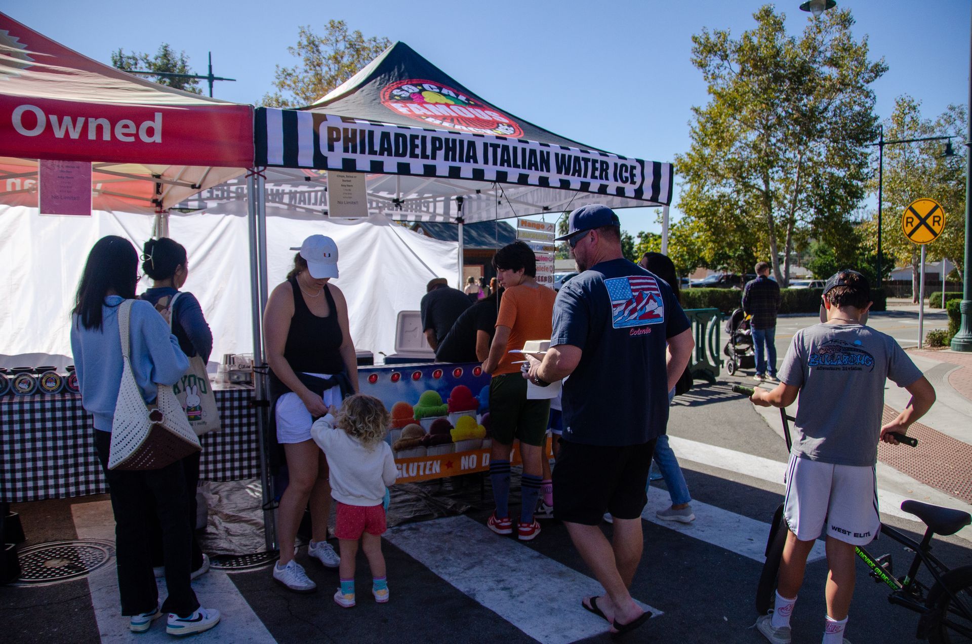 A food stall selling Philadelphia Italian water ice with customers waiting in line outdoors on a sunny day.