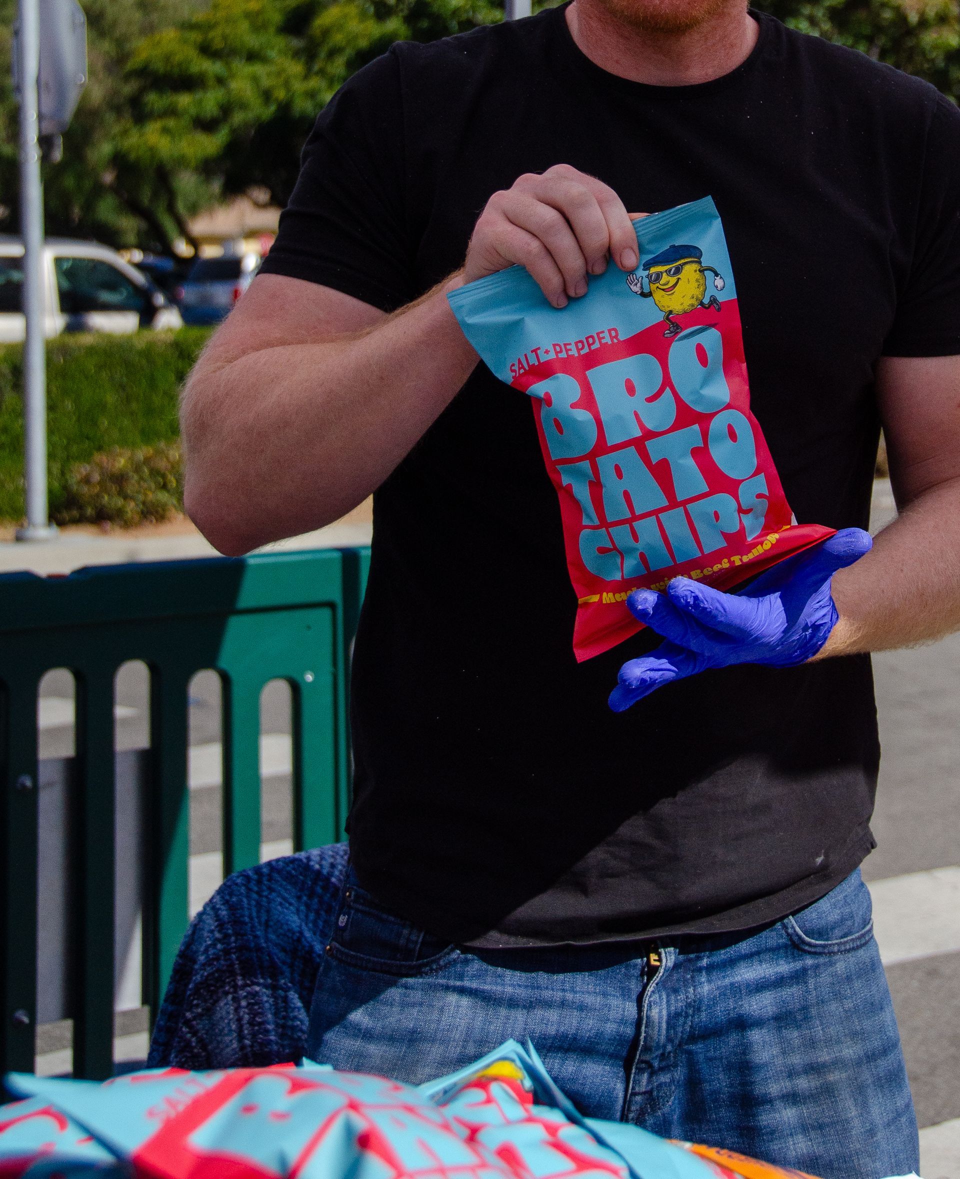 A person wearing blue gloves holds a bag of Bro Tato Chips in an outdoor setting.