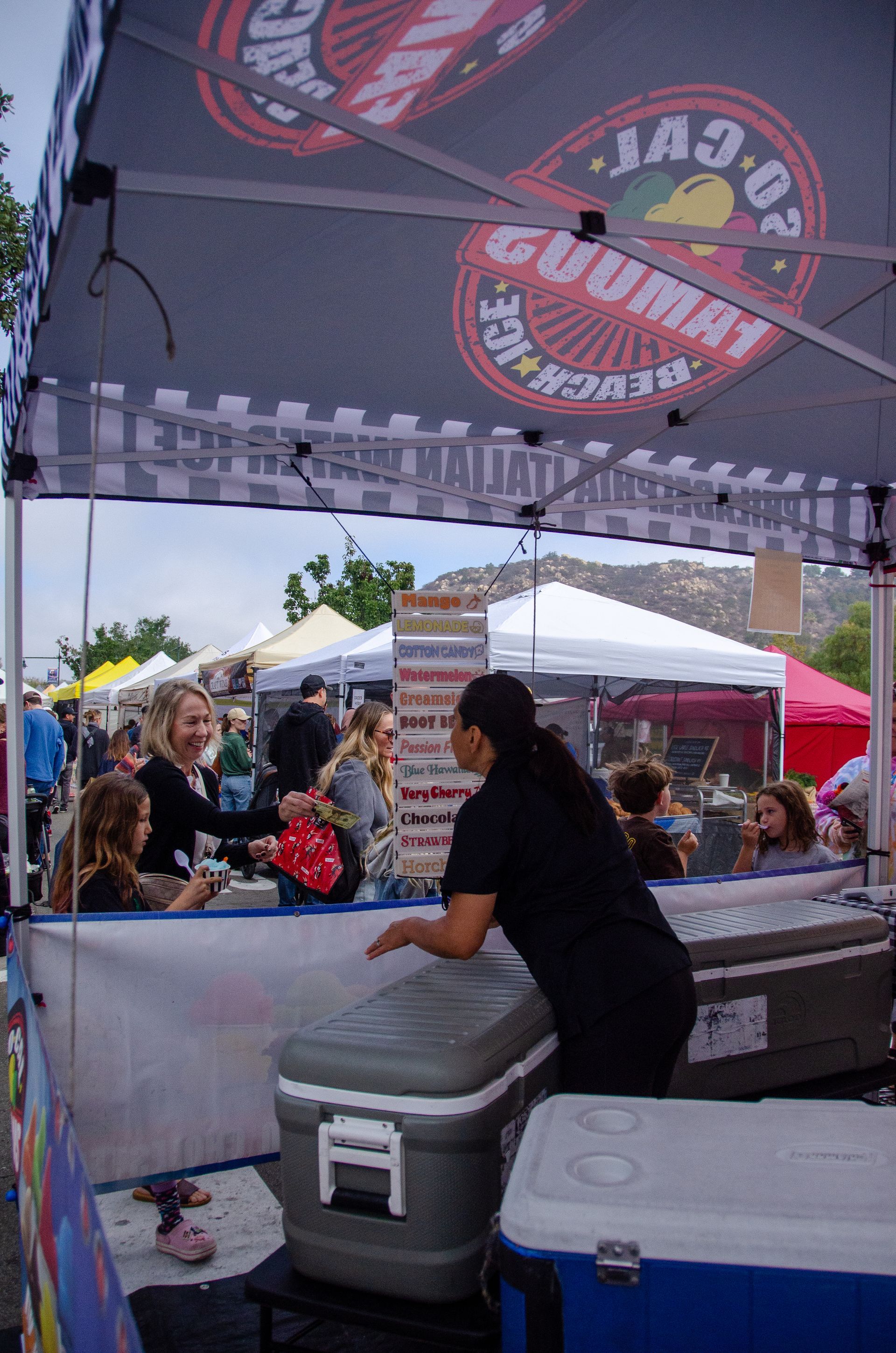 A vendor serving customers at an outdoor market stall under a branded canopy with large coolers on the counter.