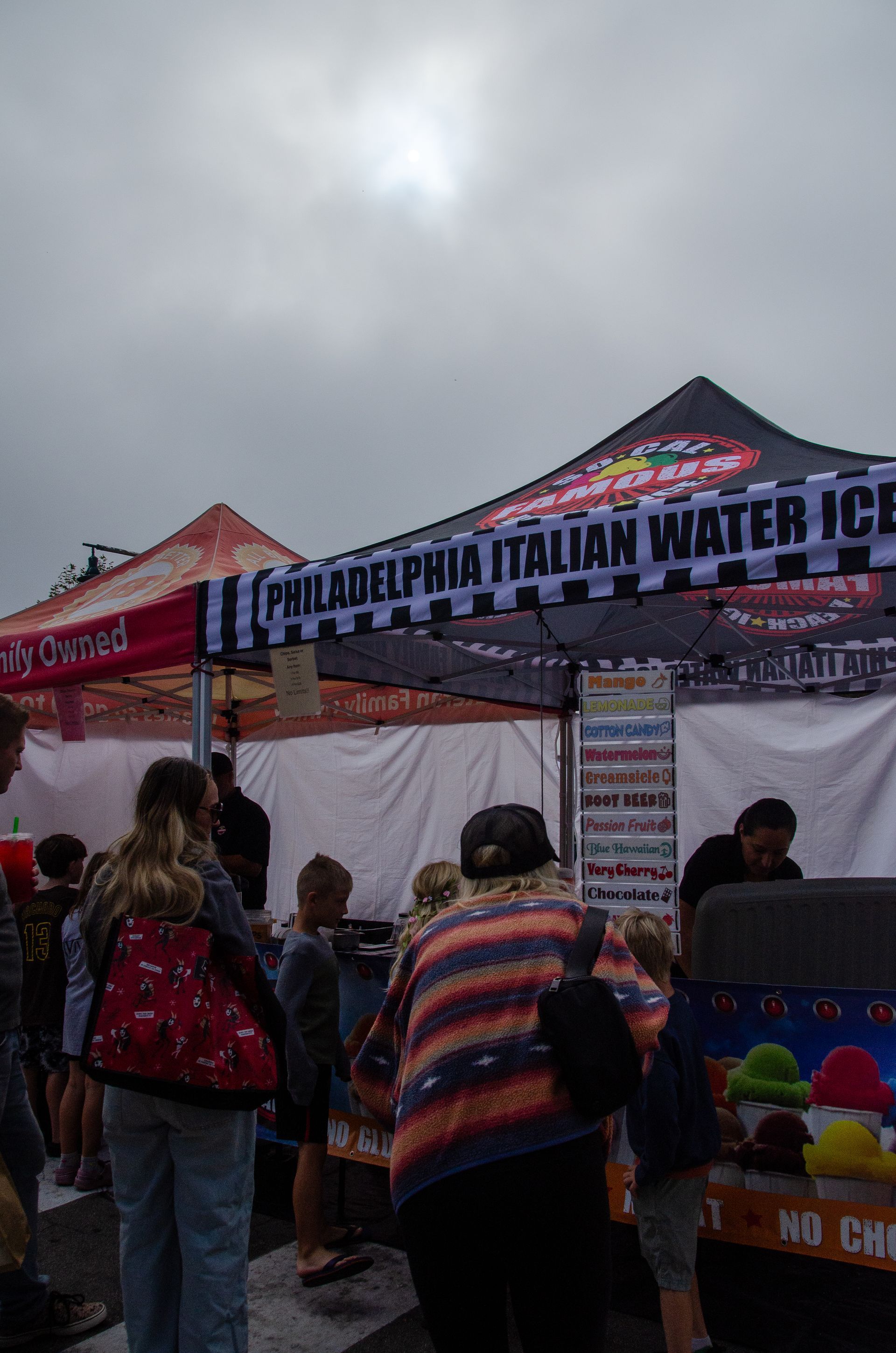 People stand in line at an outdoor Philadelphia Italian Water Ice stand under a black and white striped tent canopy.