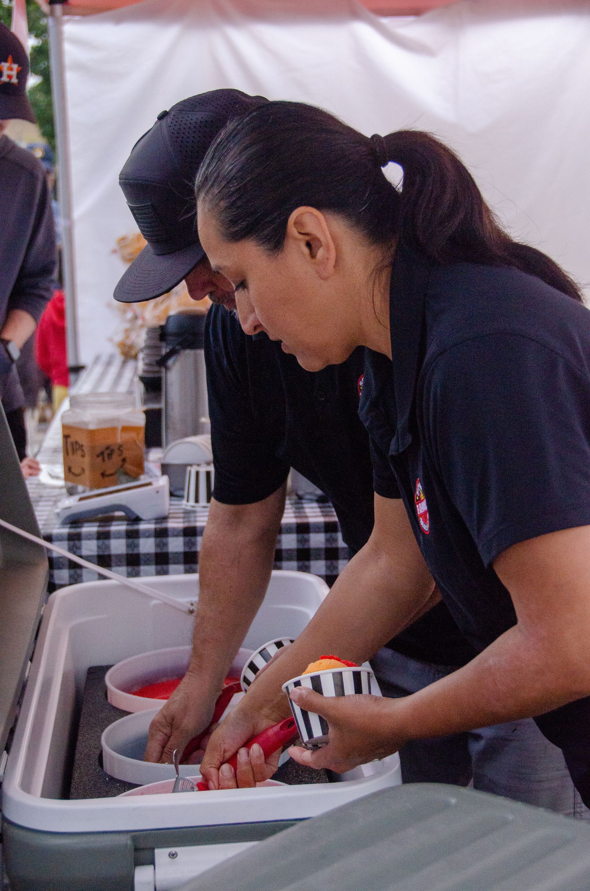 Two people wearing black shirts scoop servings into paper cups from a large white cooler at an outdoor event.