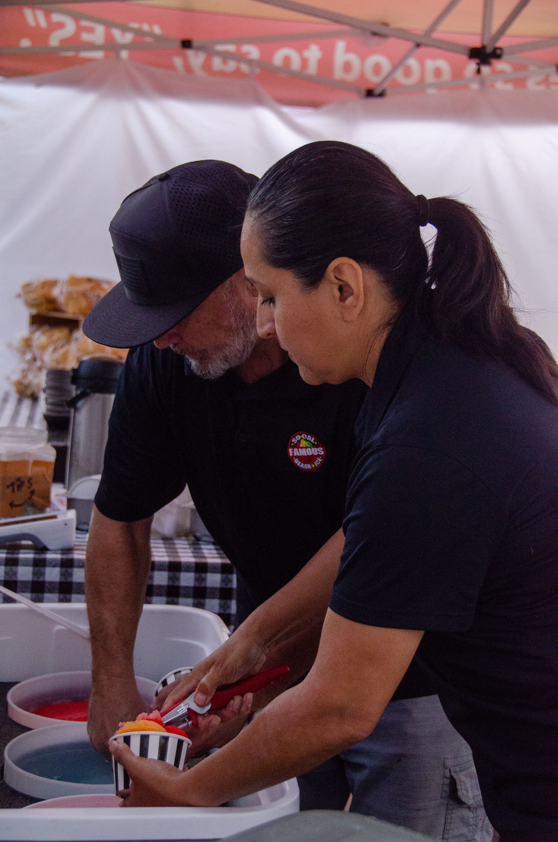 Two people in black uniforms work together at a food stall, preparing a colorful shaved ice treat in a small cup.