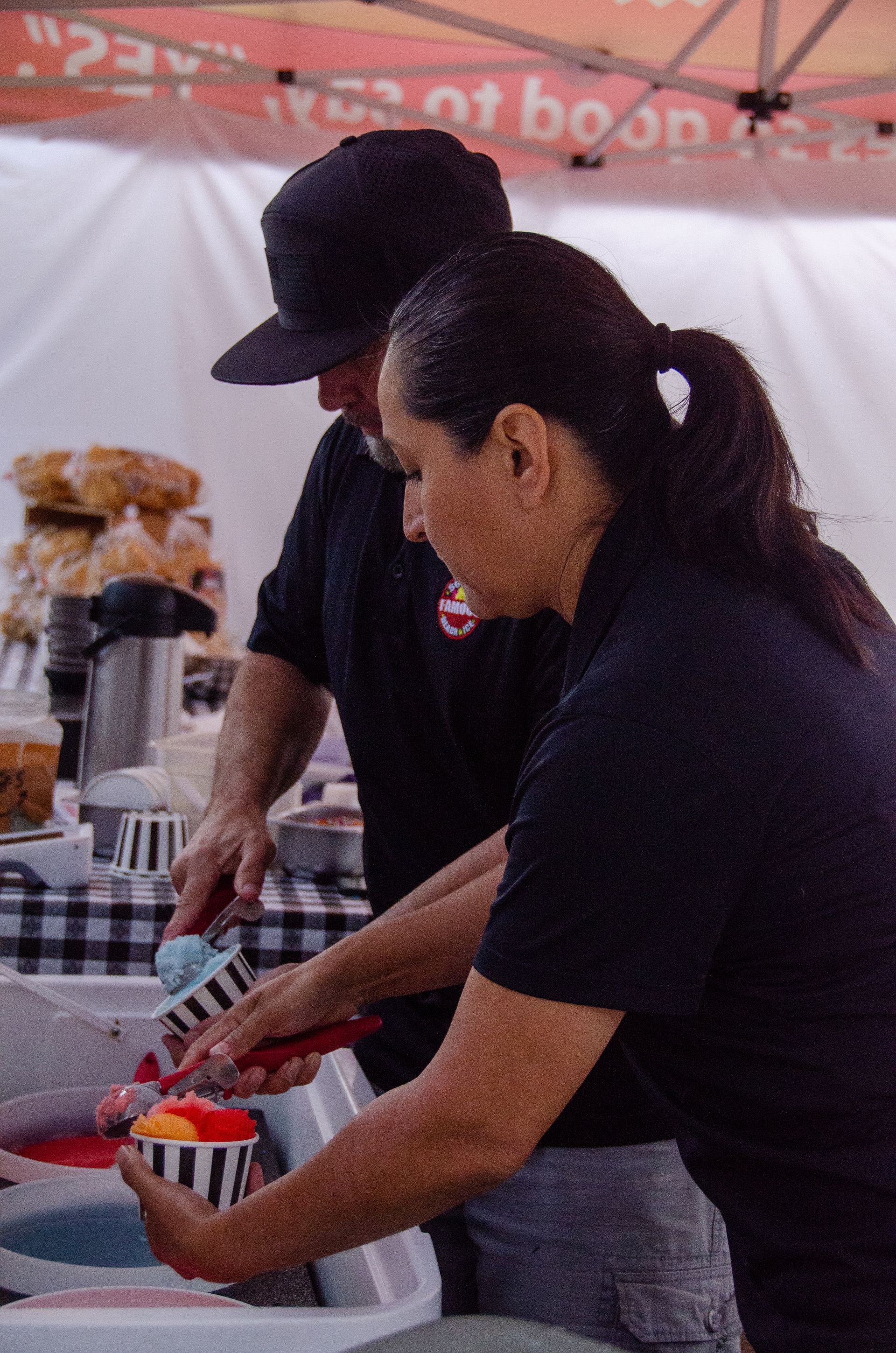 Two people in black shirts work under a tent, scooping colorful shaved ice into striped paper cups at a food stall.