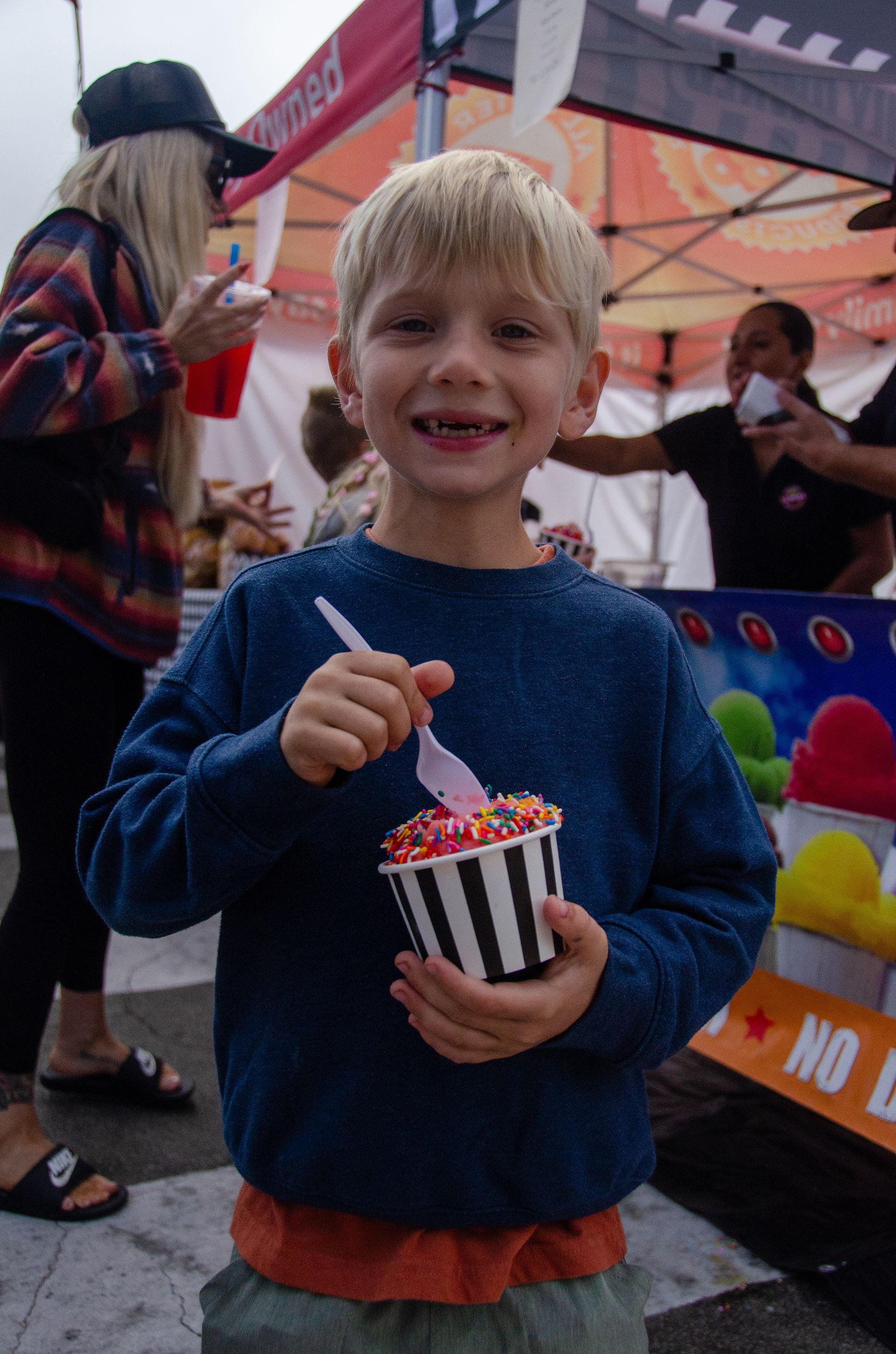 A young person in a blue sweater holds a striped cup of ice cream with sprinkles, smiling in front of a festival tent.