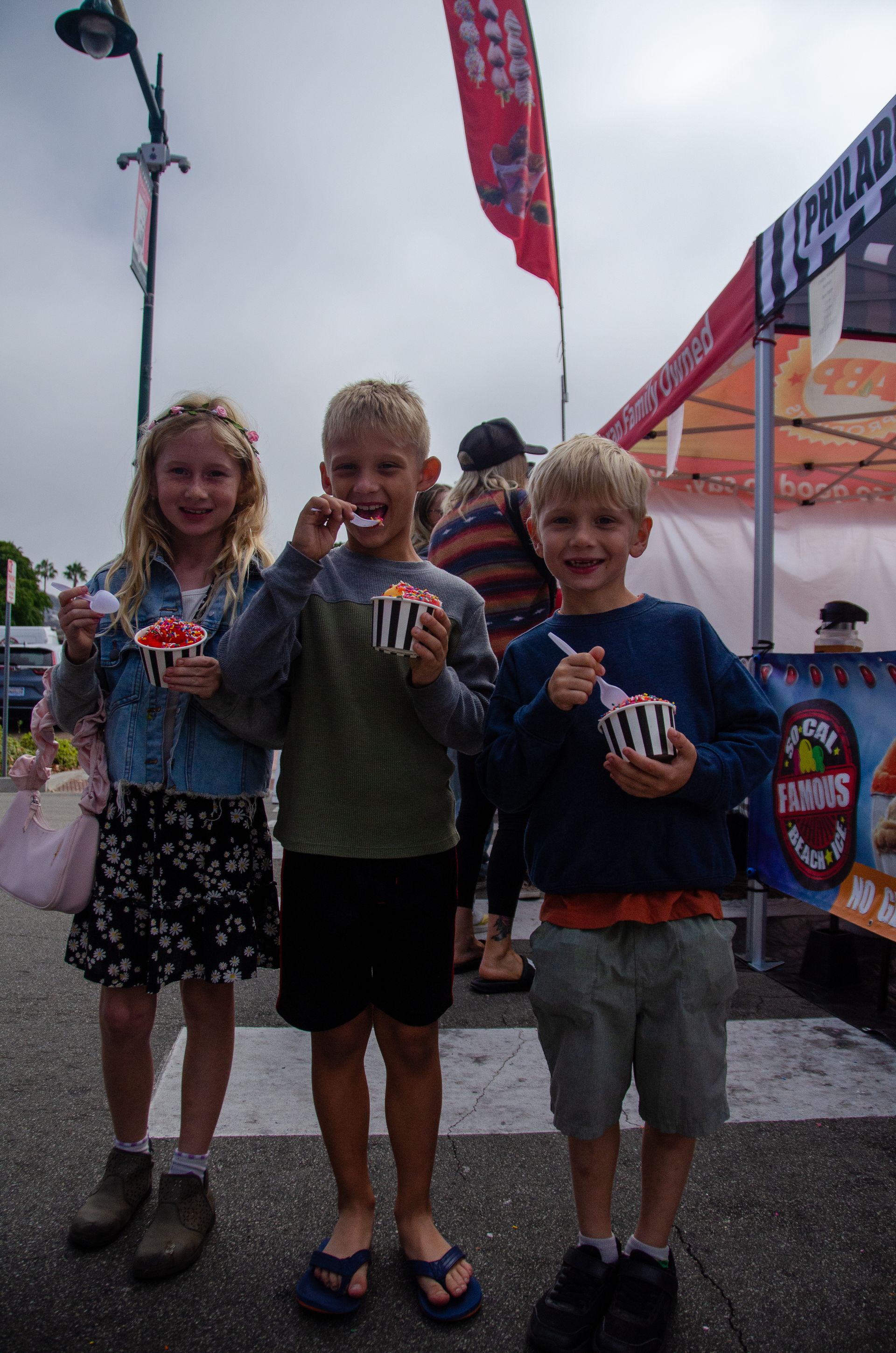 Three children holding cups of shaved ice, smiling at an outdoor fair.