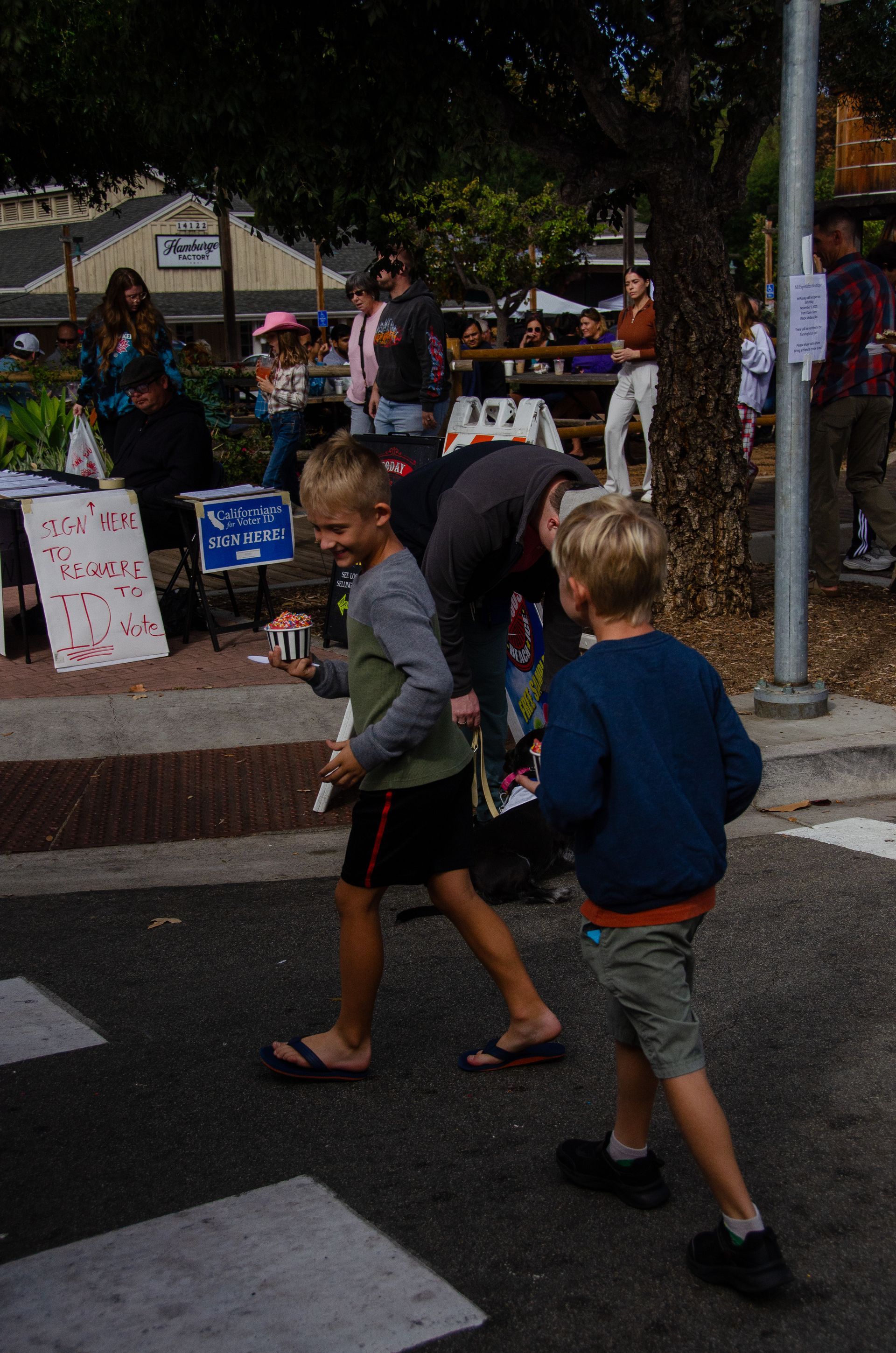 Two children walk across a crosswalk at an outdoor community event with vendors and a crowd in the background.