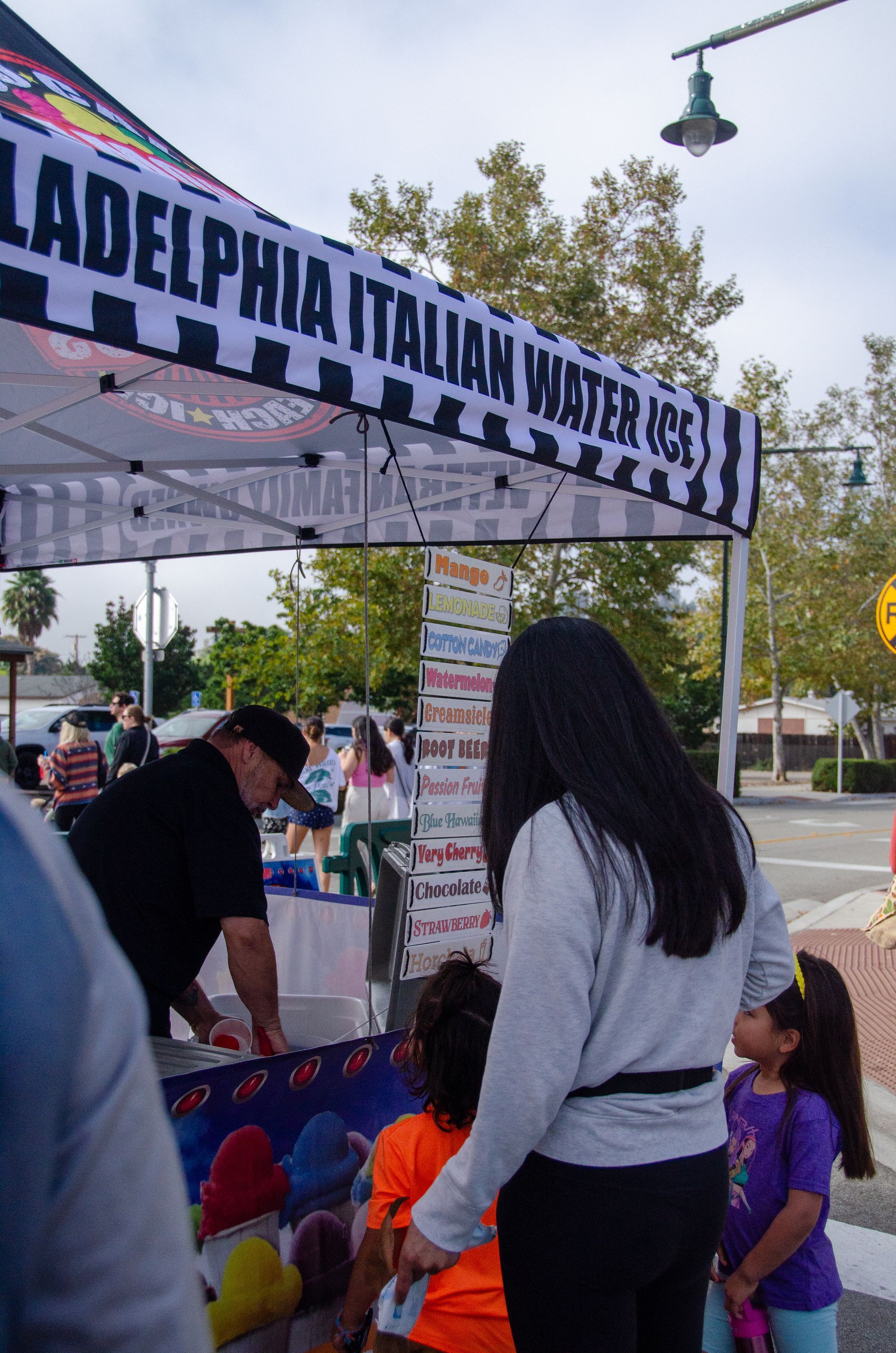 A person serves Philadelphia Italian Water Ice from a striped outdoor vendor tent to customers standing at the counter.