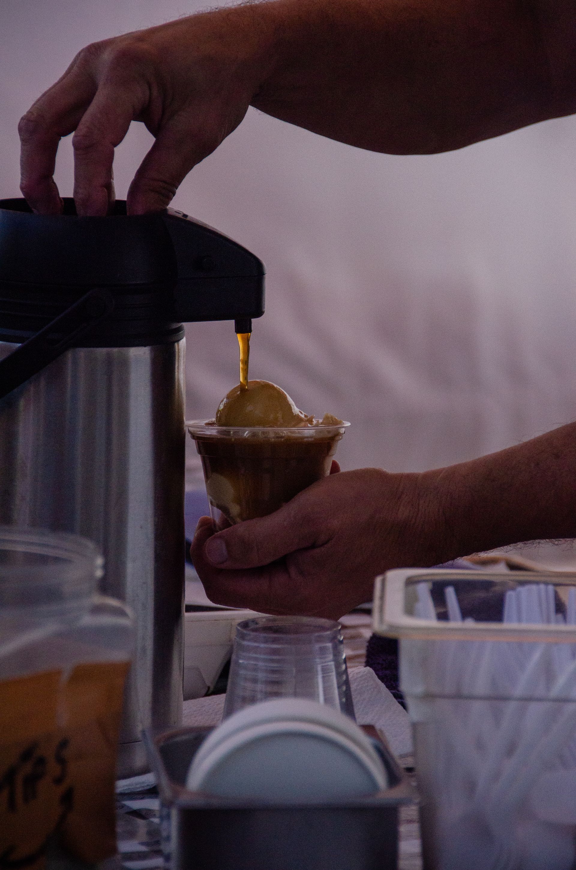 A person pours hot coffee from a carafe into a clear plastic cup filled with ice cream to make an affogato.