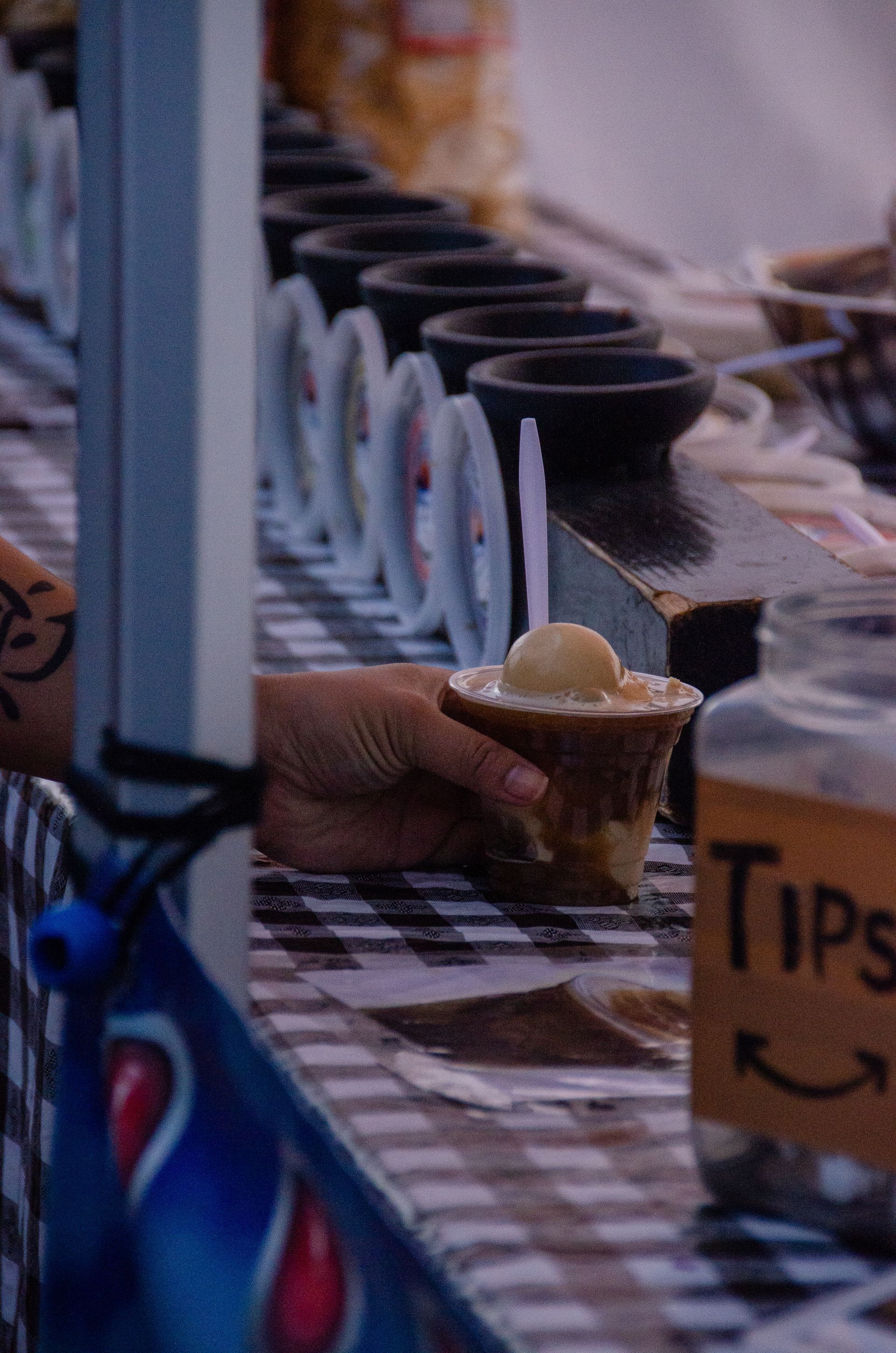 A hand holds a cup with a scoop of ice cream on a checkered table next to a tip jar at an outdoor food stand.