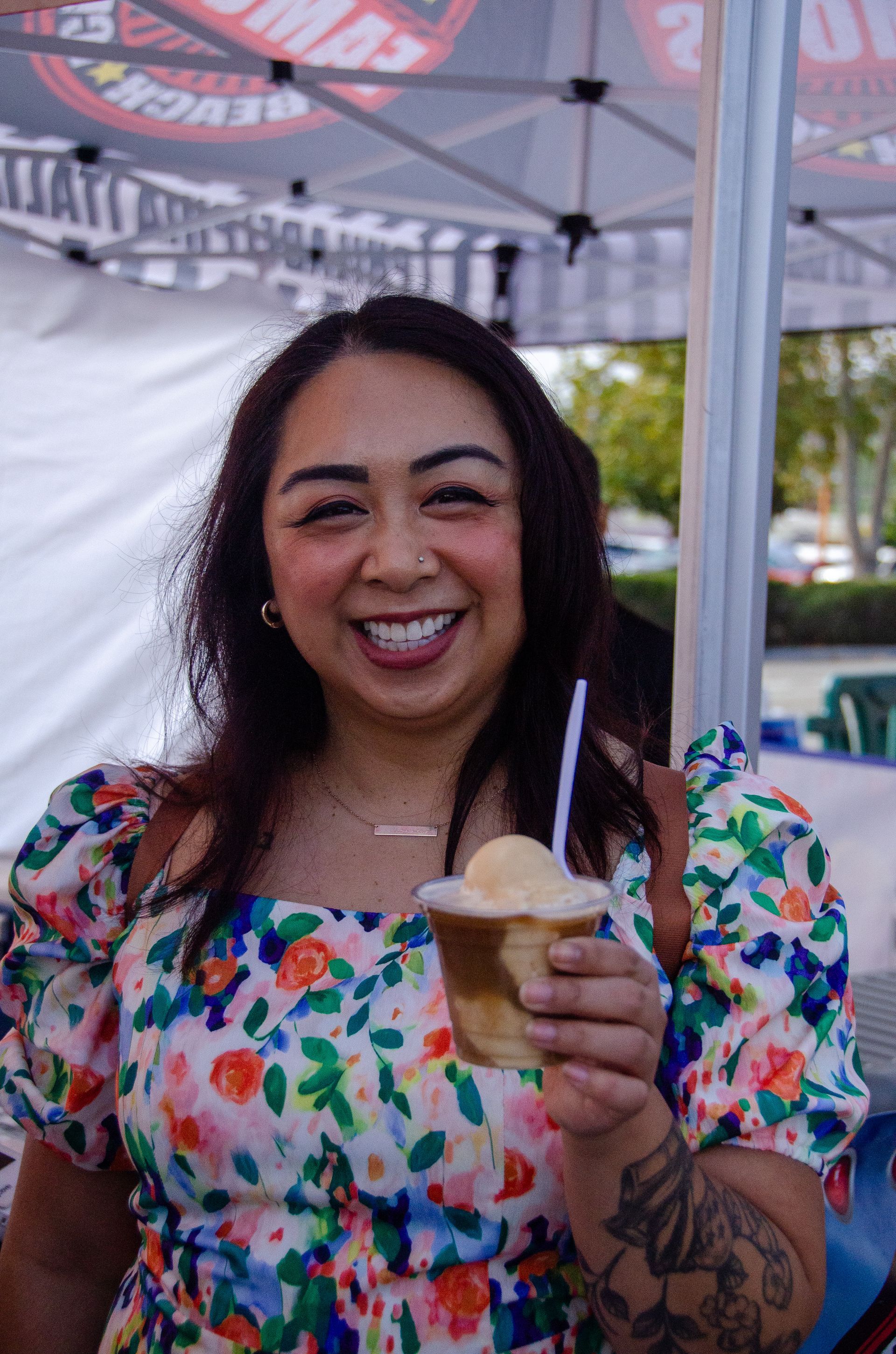 Smiling person holding a dessert cup with a scoop of ice cream at an outdoor event under a branded canopy.