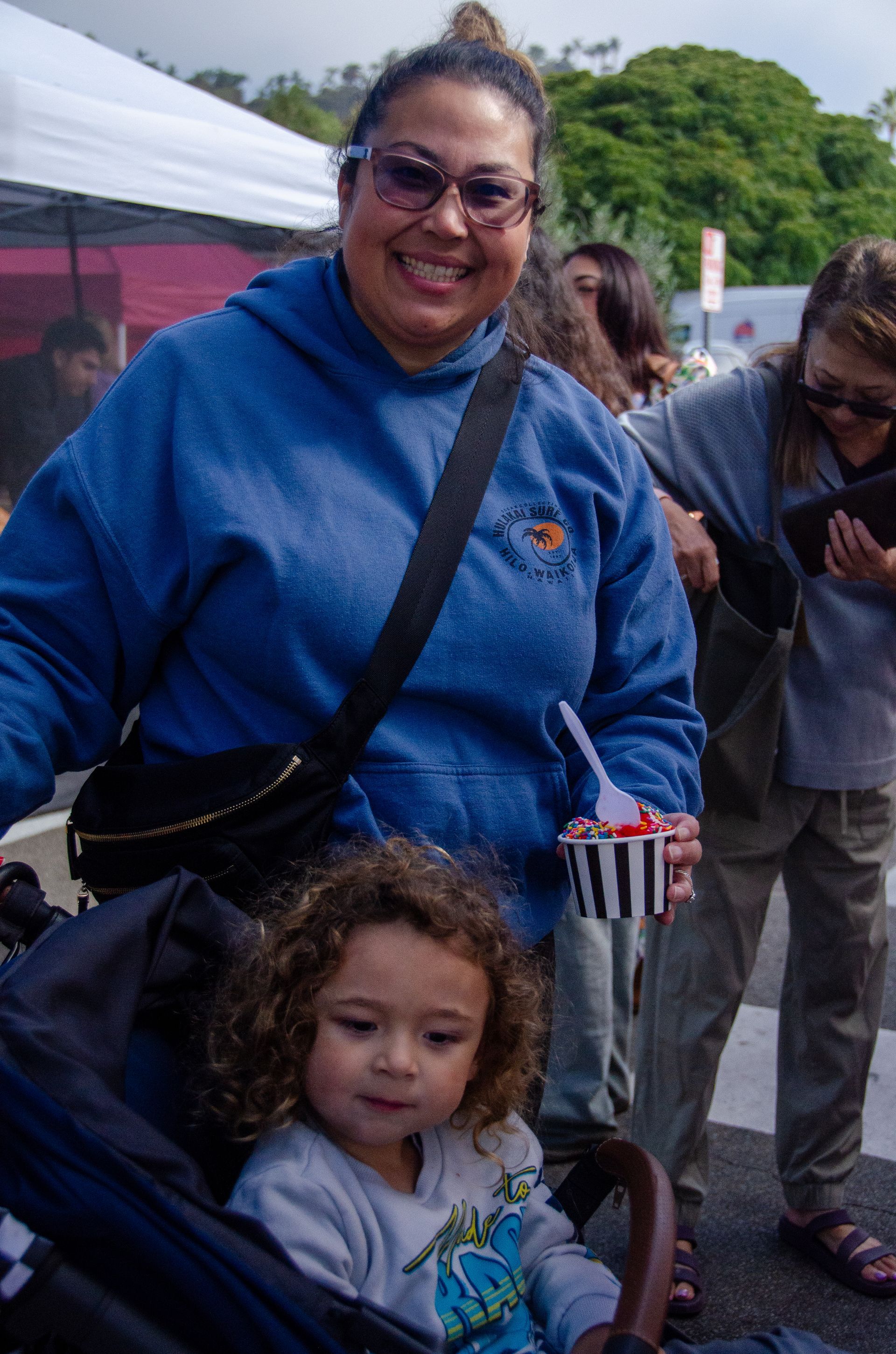 A smiling person in a blue hoodie holds a striped cup with a spoon, next to a child in a stroller outdoors.