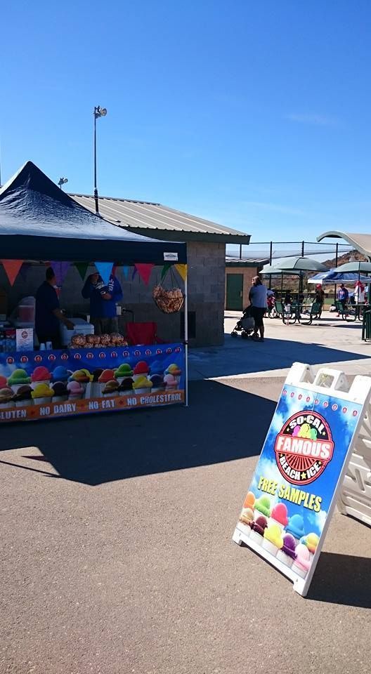 A snow cone vendor stand with a blue canopy and triangular flags under a clear blue sky, featuring a branded sign.