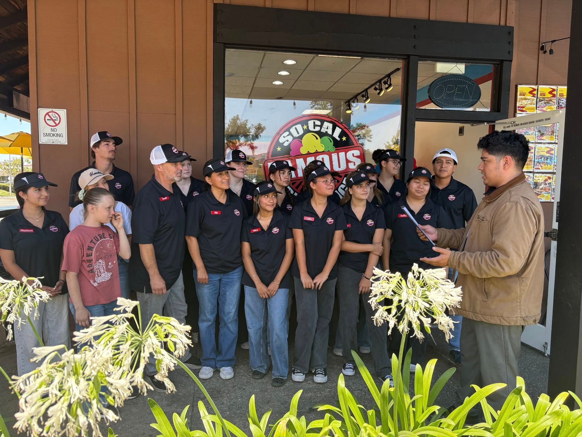 A group of employees in black shirts and hats stand outside a restaurant, listening to a man in a tan jacket speaking.