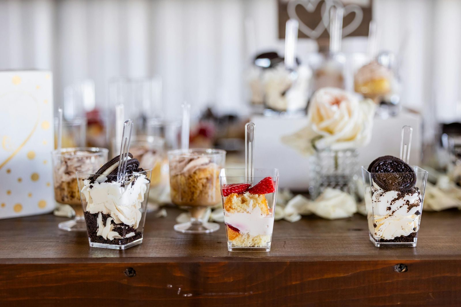 A wooden table displays several small, individual dessert cups with treats like layered cream, cookies, and strawberries.