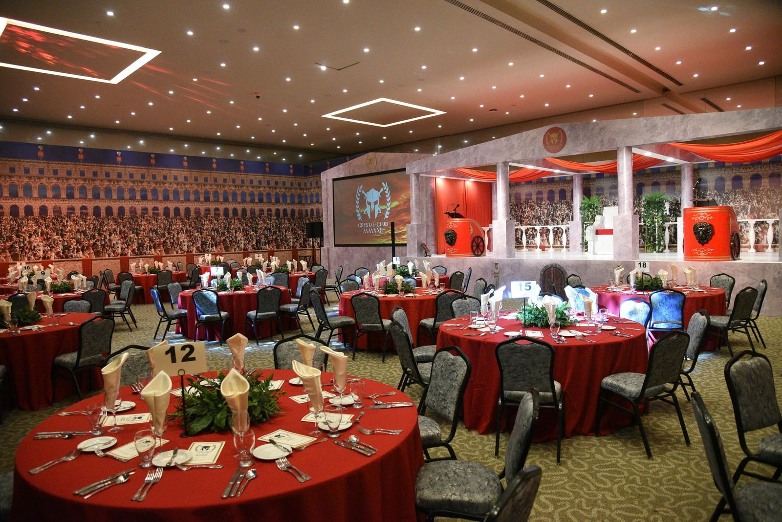 A ballroom set for a formal event with red-clothed round tables, place settings, and a stage with classical architecture.