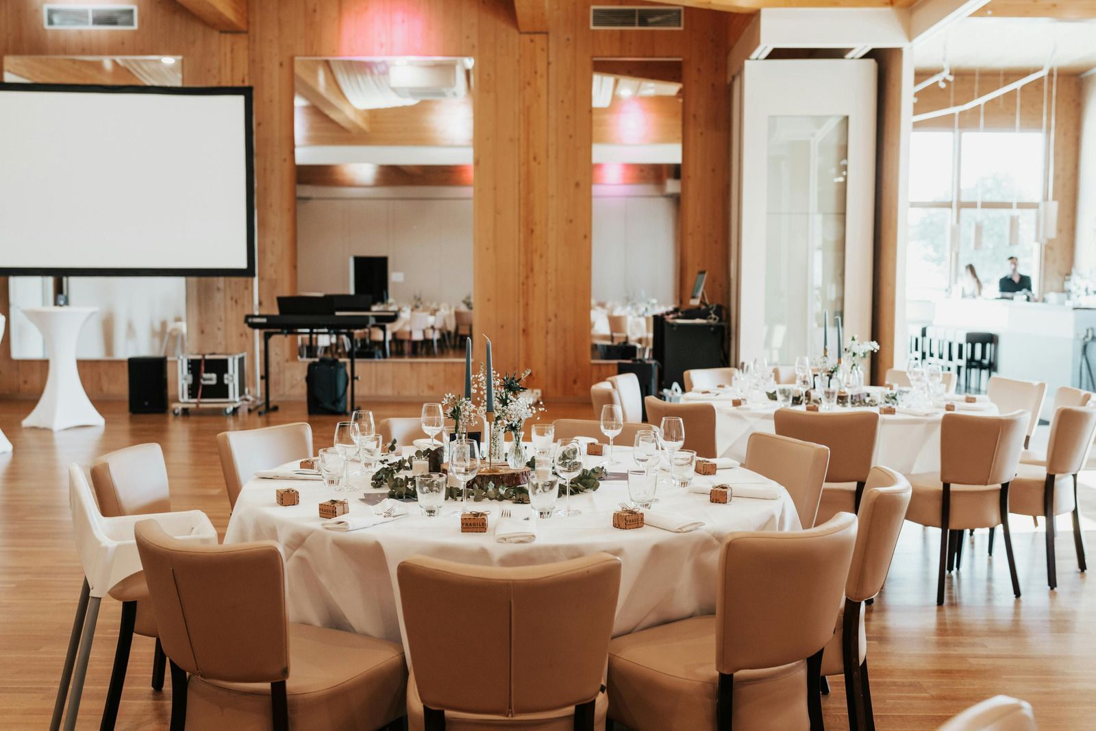 A wedding reception setup with round tables, tan chairs, and floral centerpieces in a wood-paneled banquet hall.