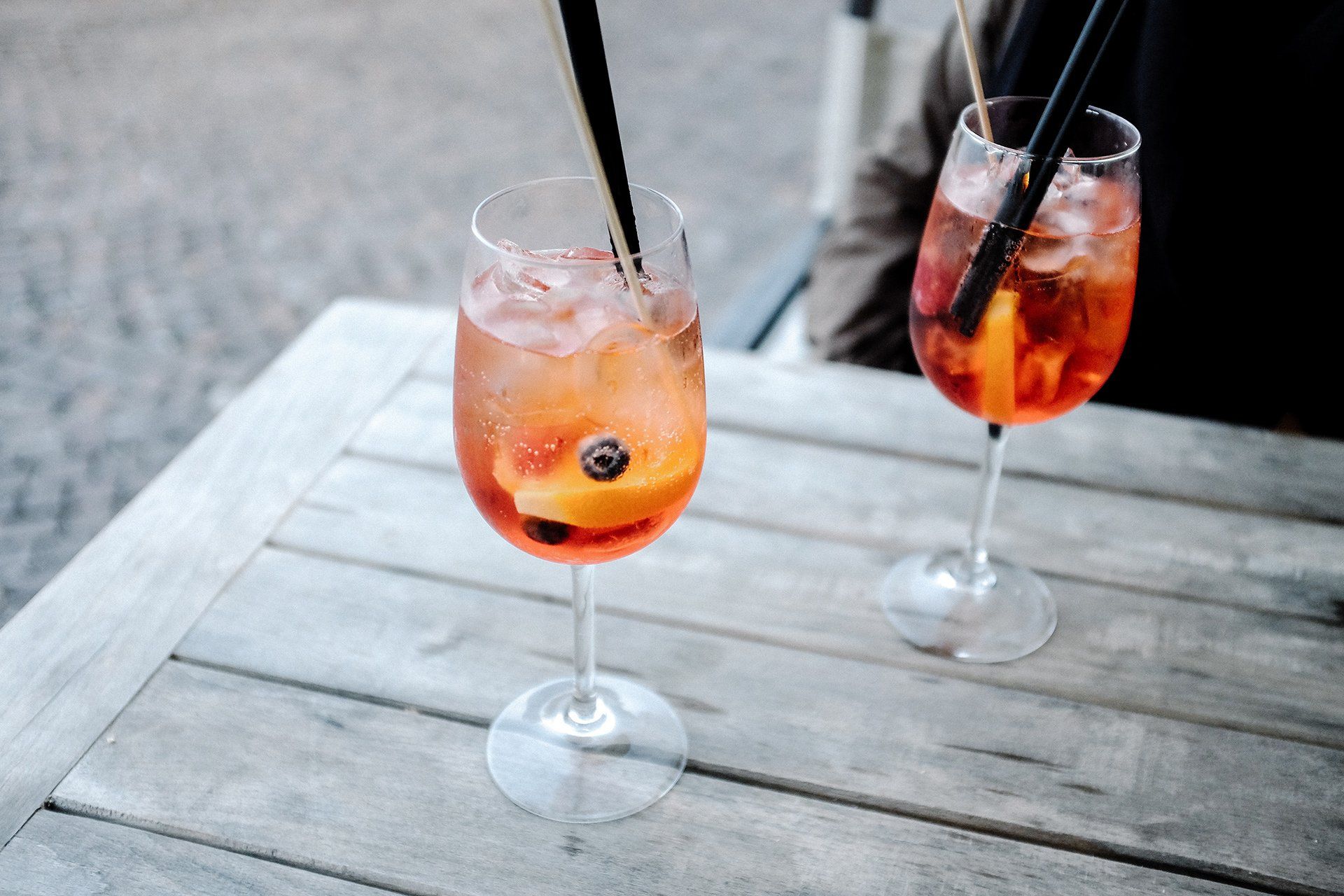 Two glasses of orange cocktails with ice and fruit slices sit on a weathered wooden table outdoors.