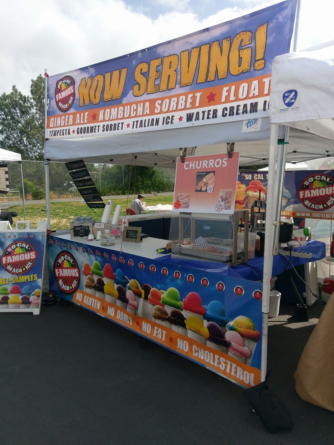A blue food stall tent under a blue sky, advertising churros, kombucha, sorbet, and floats, with a sign listing menu items.