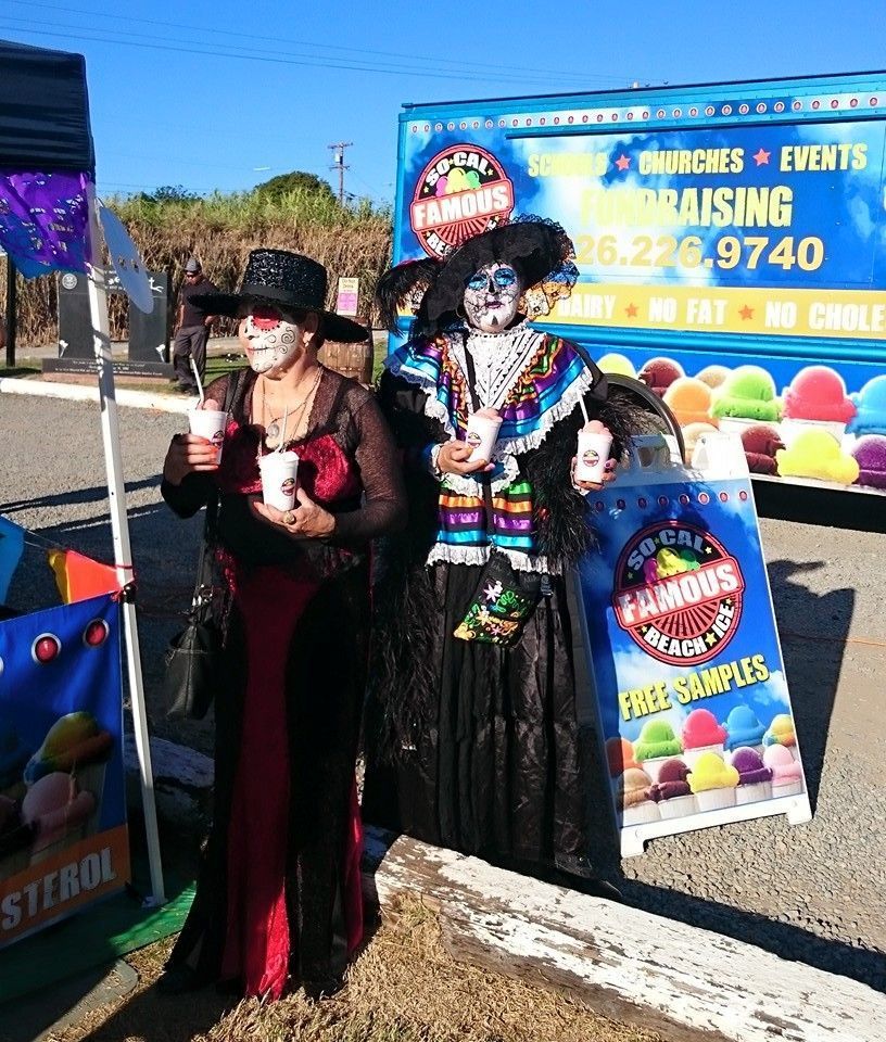 Two people in Day of the Dead costumes and makeup stand outside by a shaved ice truck, holding cups of shaved ice.