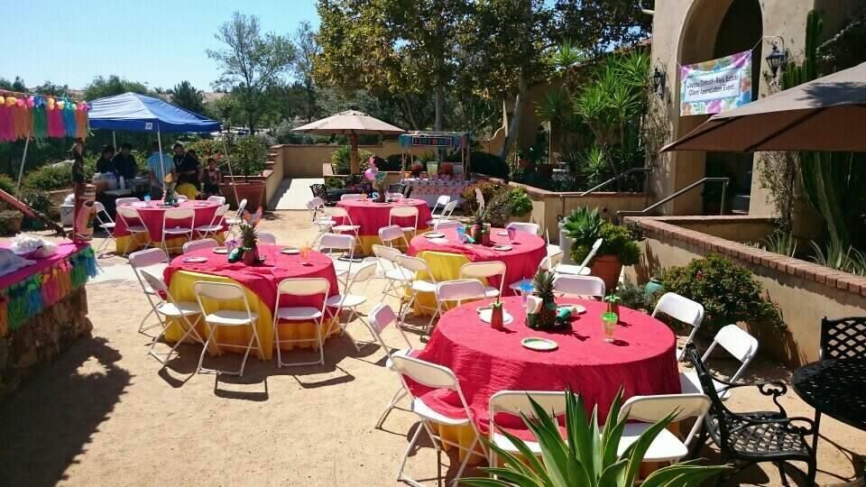 An outdoor patio area with multiple round tables covered in red and yellow tablecloths, set for an event with white chairs.