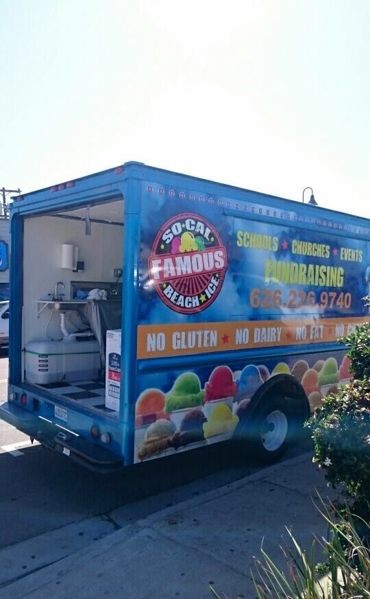 A blue So-Cal Famous Beach Ice food truck parked on a street, featuring colorful shaved ice graphics and phone number.