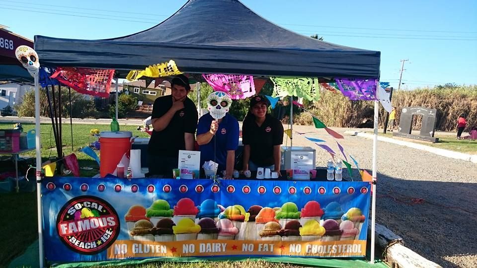 Three people staff a food stall with a banner displaying colorful shaved ice, decorated with festive skull ornaments.