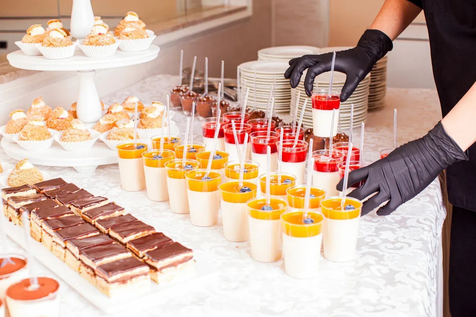 A person wearing black gloves sets a small dessert cup on a table filled with various treats, including layered desserts.