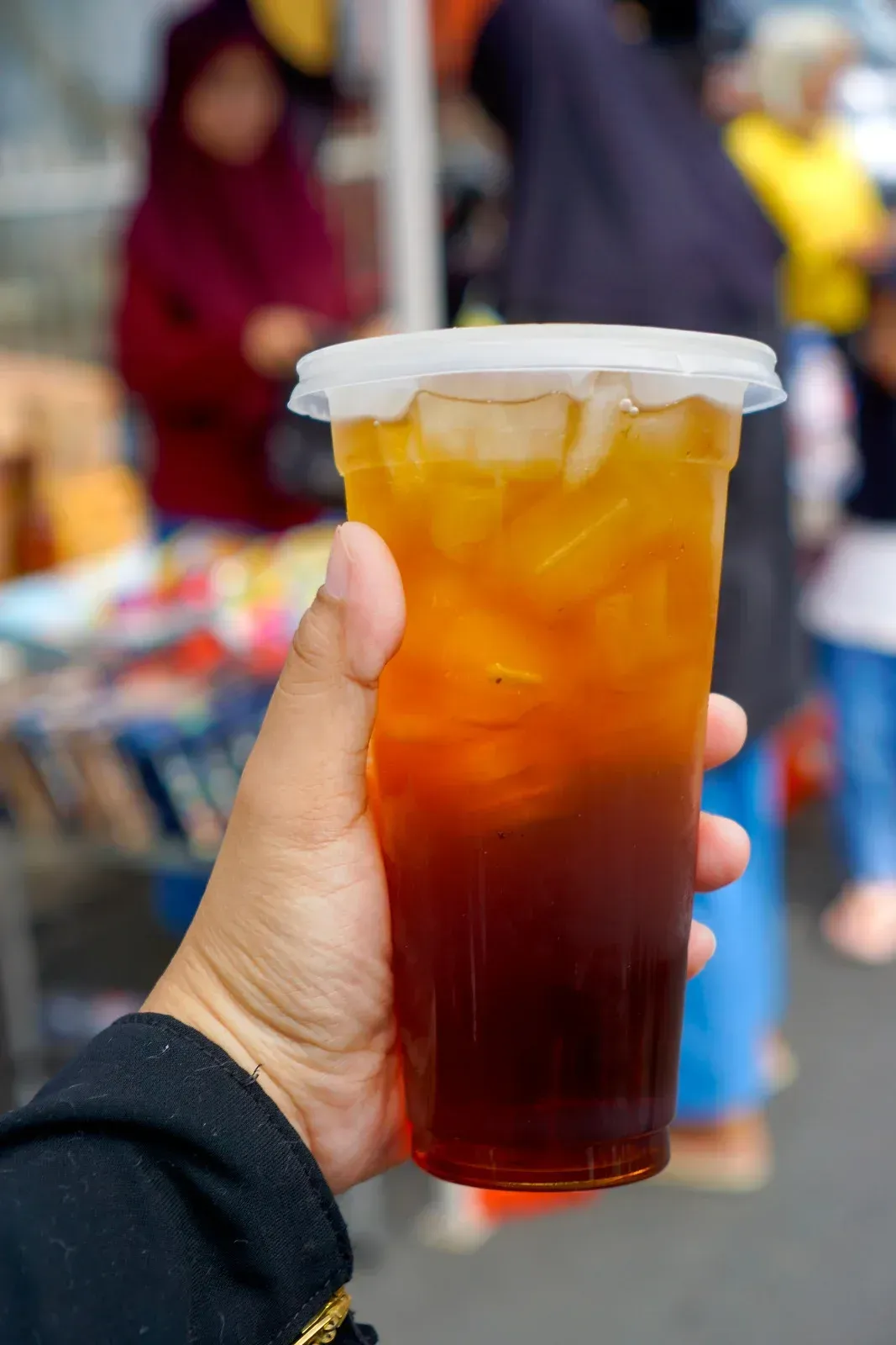 A hand holds a plastic cup of iced tea with a gradient of amber to dark brown liquid against a blurred outdoor market.