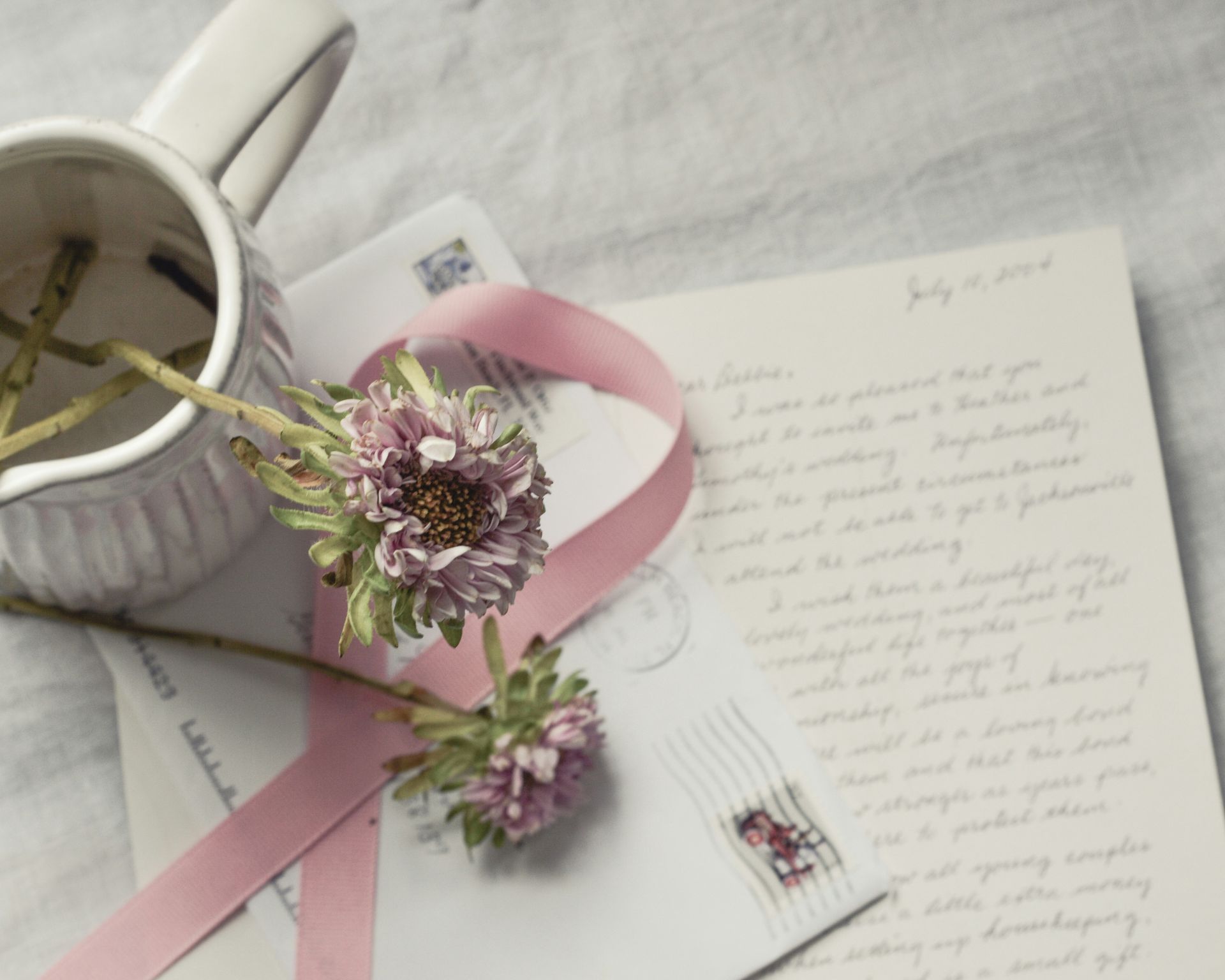 Flowers, a pink ribbon, and handwritten notes beside a cup on a soft white surface