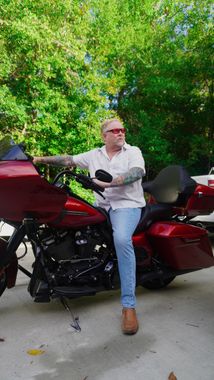 Man with sunglasses sits on red motorcycle, arm outstretched. Outdoors with trees in the background.