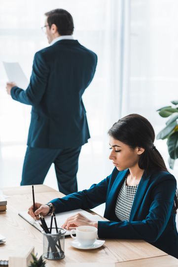 Woman in blue suit writes at a desk; man in a suit looks at papers in the background. Office setting.