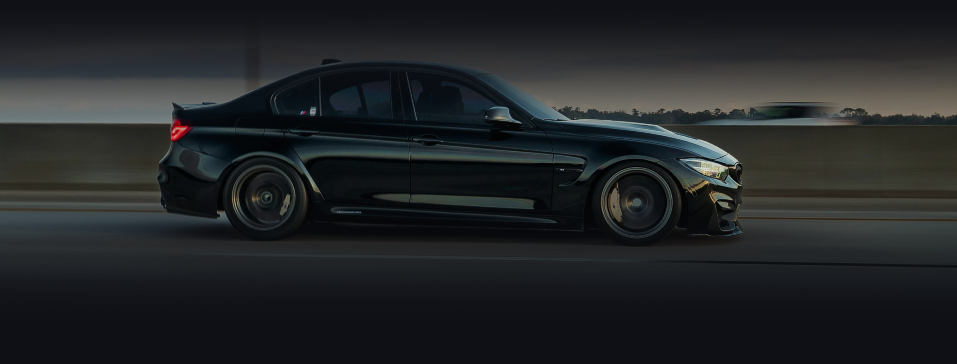 A sleek black BMW M3 driving at speed on a highway, shown from a side profile with motion-blurred wheels.