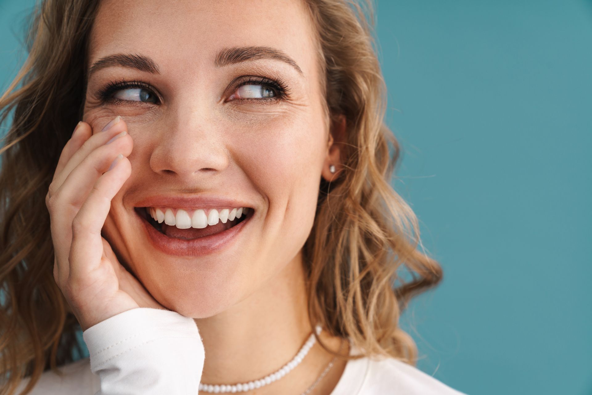 Happy woman with bright white teeth and smooth complexion smiling confidently in a blue background, showing off the results of veneers in Newtown.
