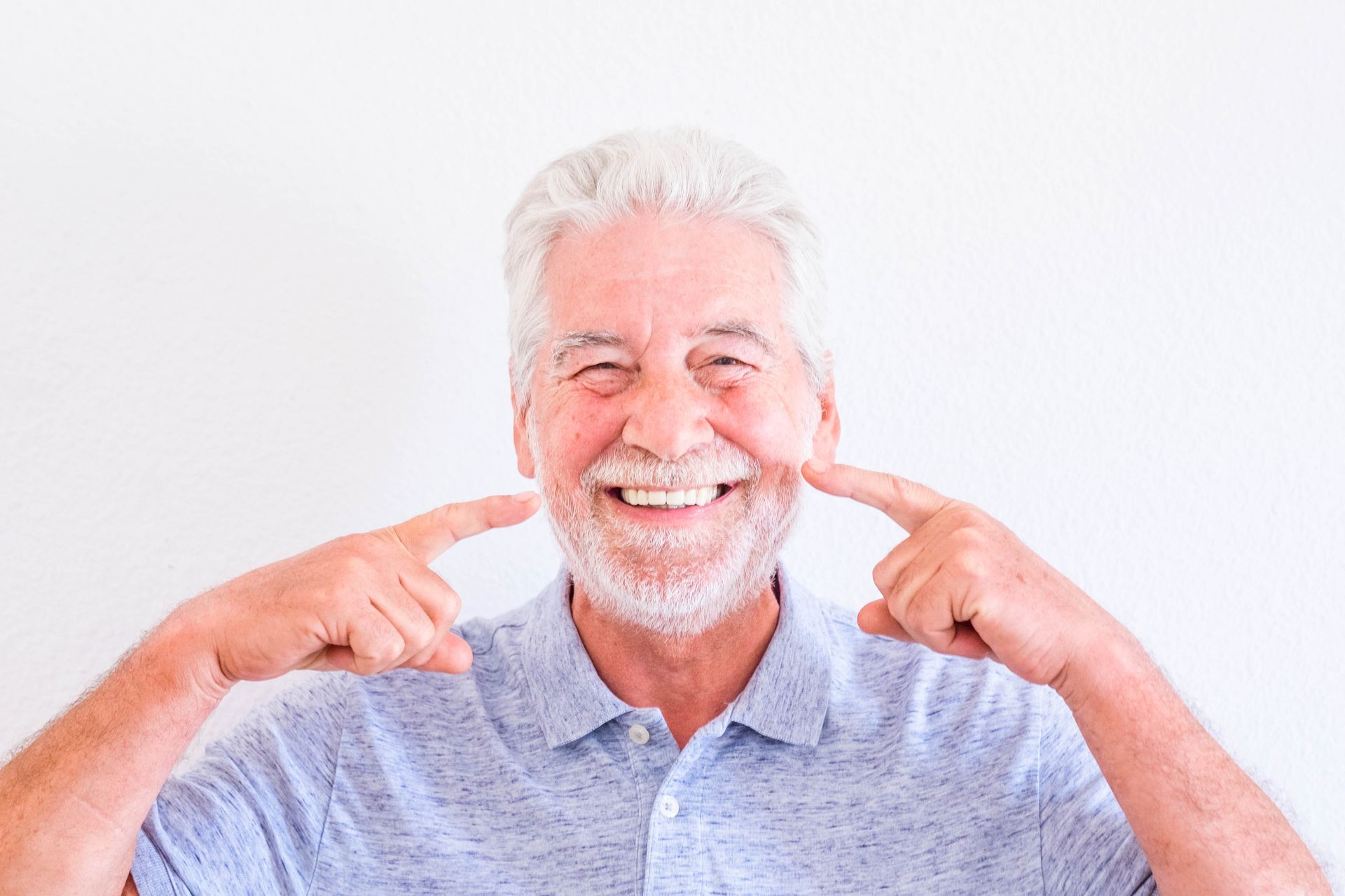 Confident senior man with white hair and beard smiling brightly and pointing to his teeth after receiving All-on-4 dental implants in Toowoomba, showcasing a full set of natural-looking teeth at Garden City Dental Practice.