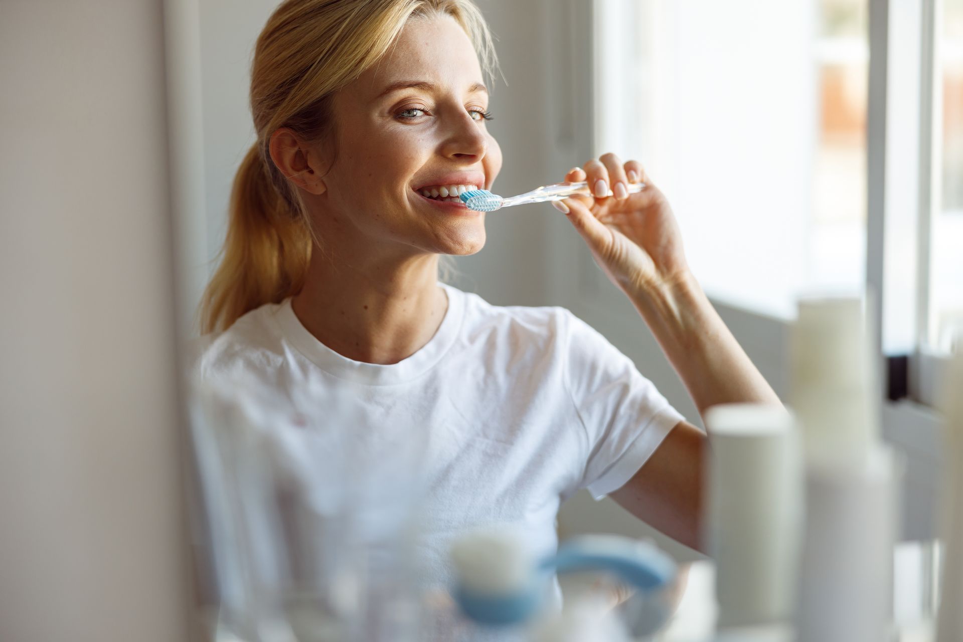 Smiling woman brushing her teeth in front of a bathroom mirror during her morning oral hygiene routine.
